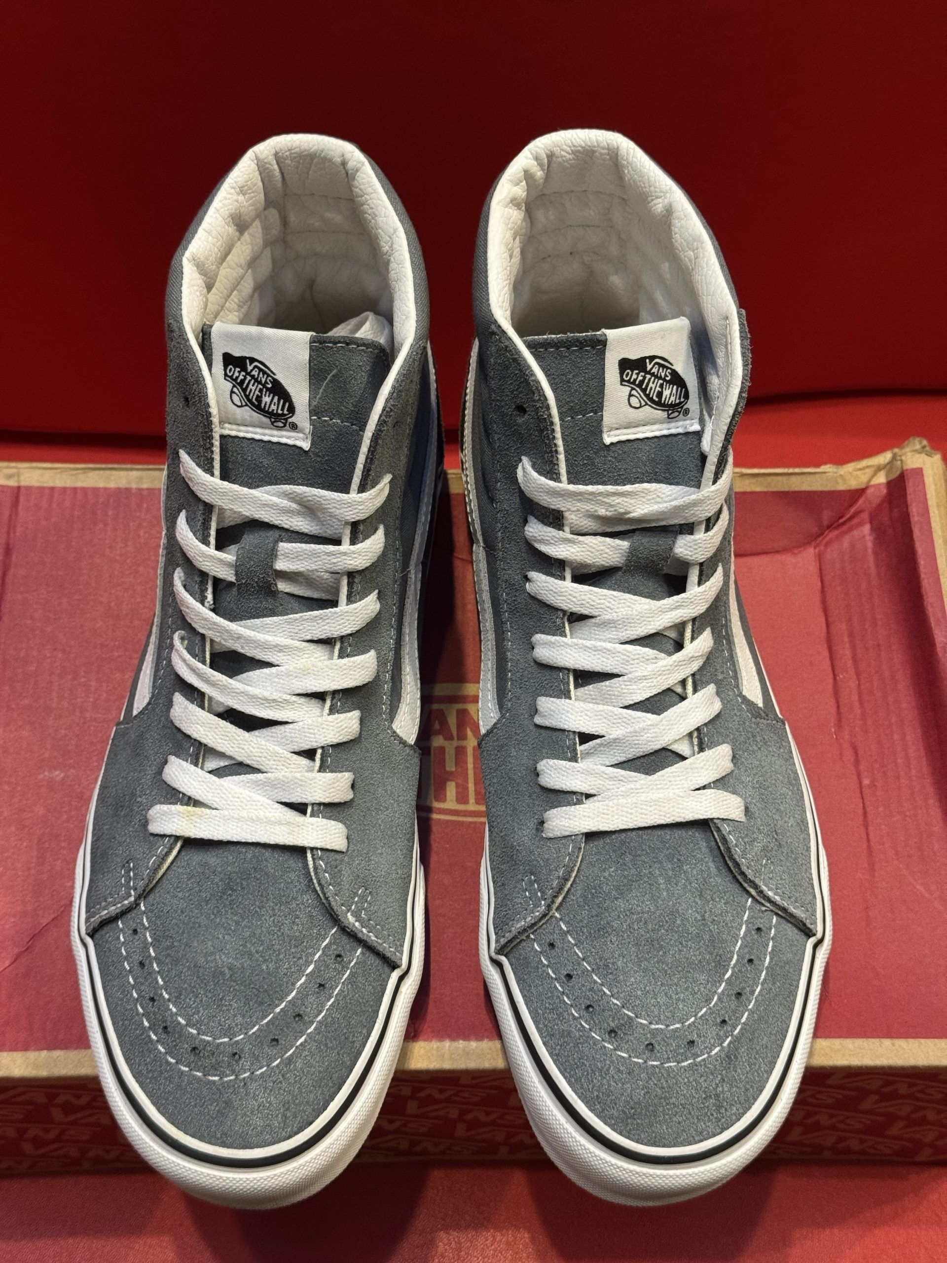 Gray Vans Sk8-Hi sneakers on a shoe box with a red background. The sneakers have white accents and a gum sole.