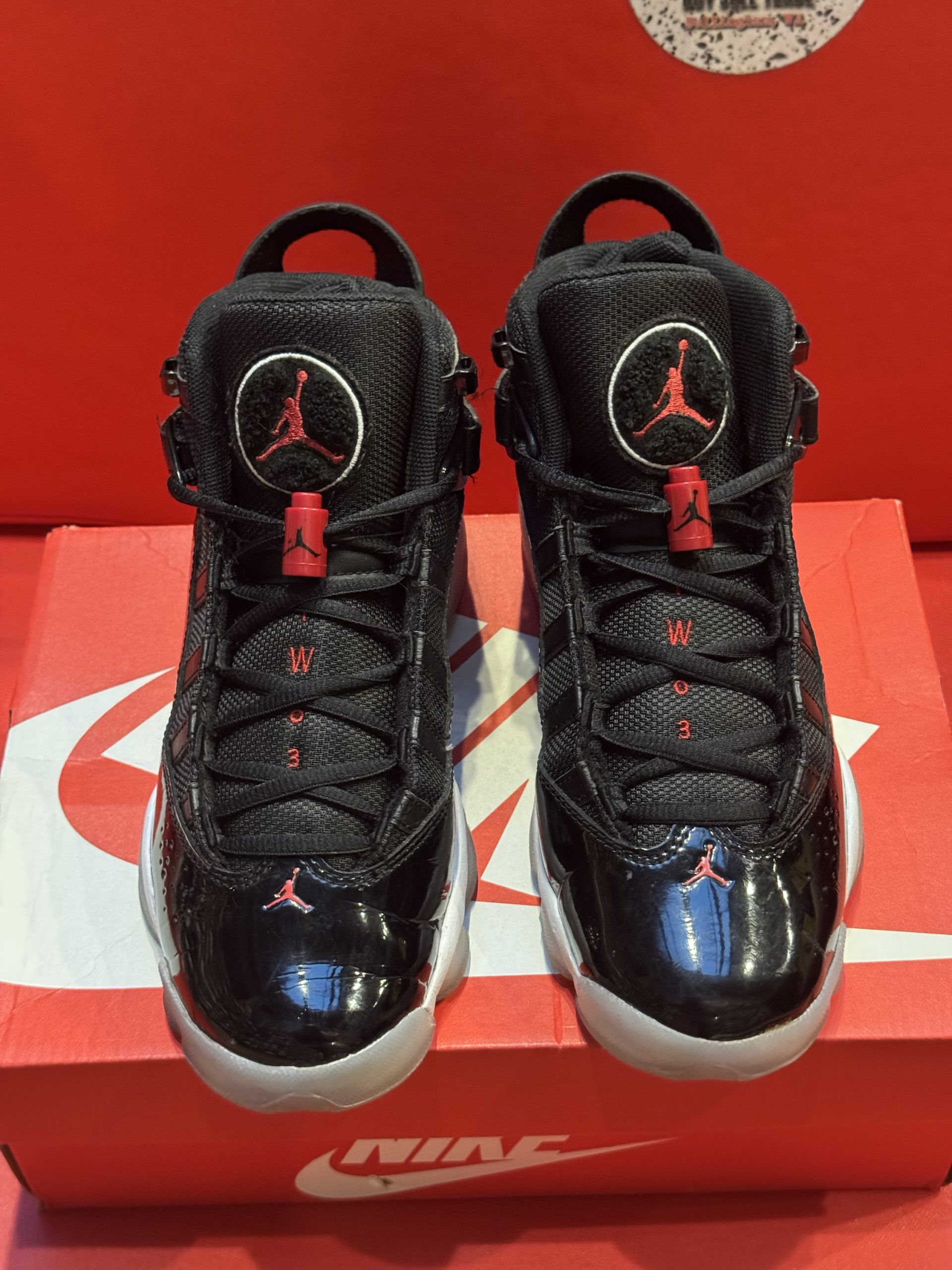Black and red Air Jordan 6 Rings sneakers on a Nike box, set against a red background.
