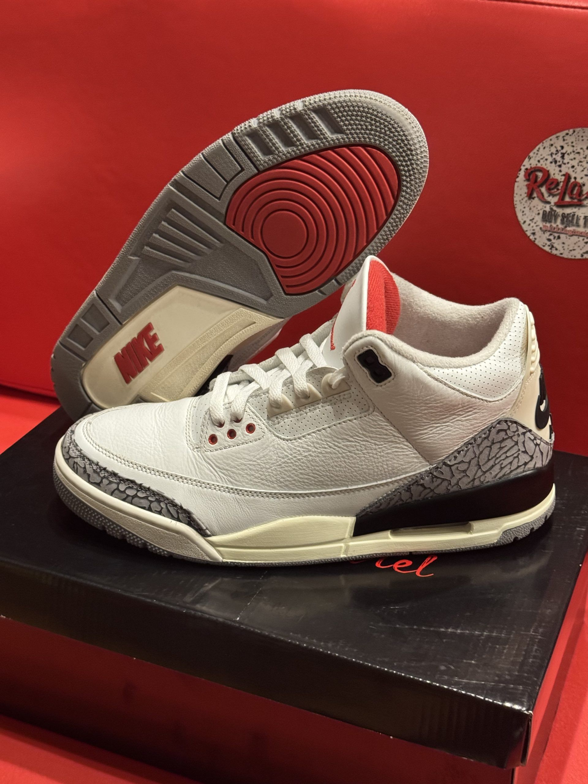 Pair of white, gray, and red Air Jordan 3 sneakers on a black box, sole up showing red details.