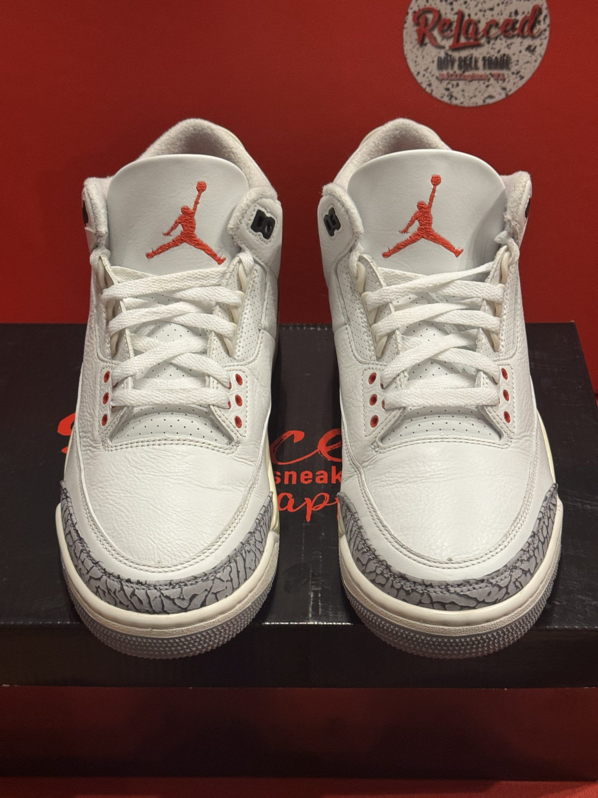 Pair of white, gray, and red Air Jordan 3 sneakers on a black box, sole up showing red details.