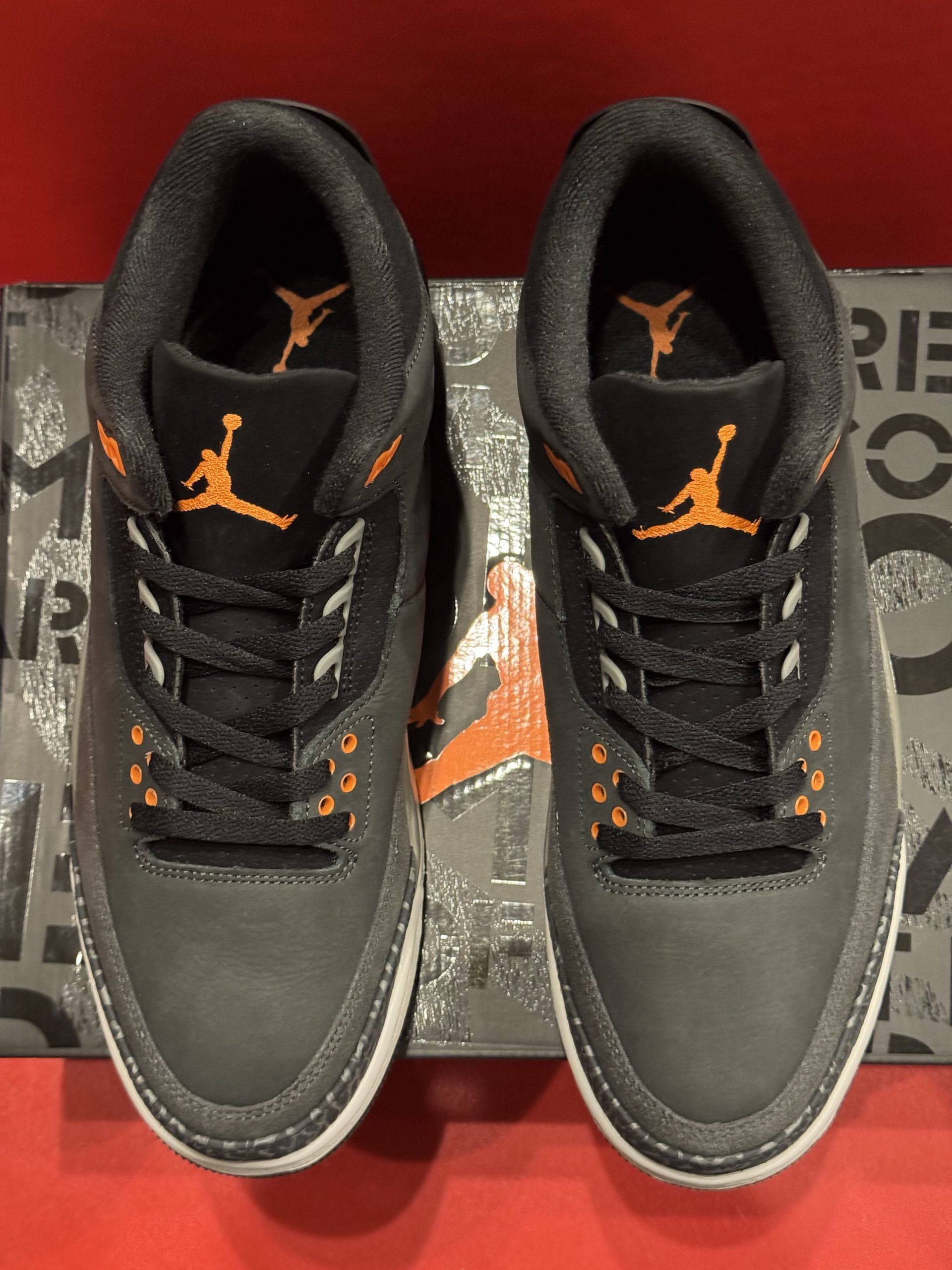 Black and orange Air Jordan 3 sneakers on a patterned box. Sole is visible, featuring orange accents.