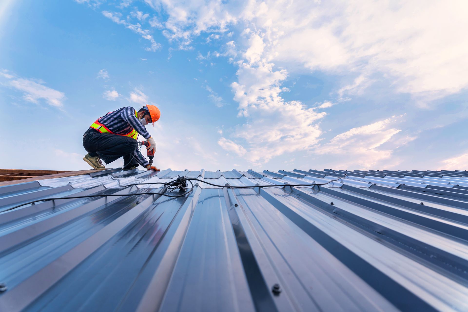 Roofer in orange helmet, securing metal roof panels against a blue sky with clouds.