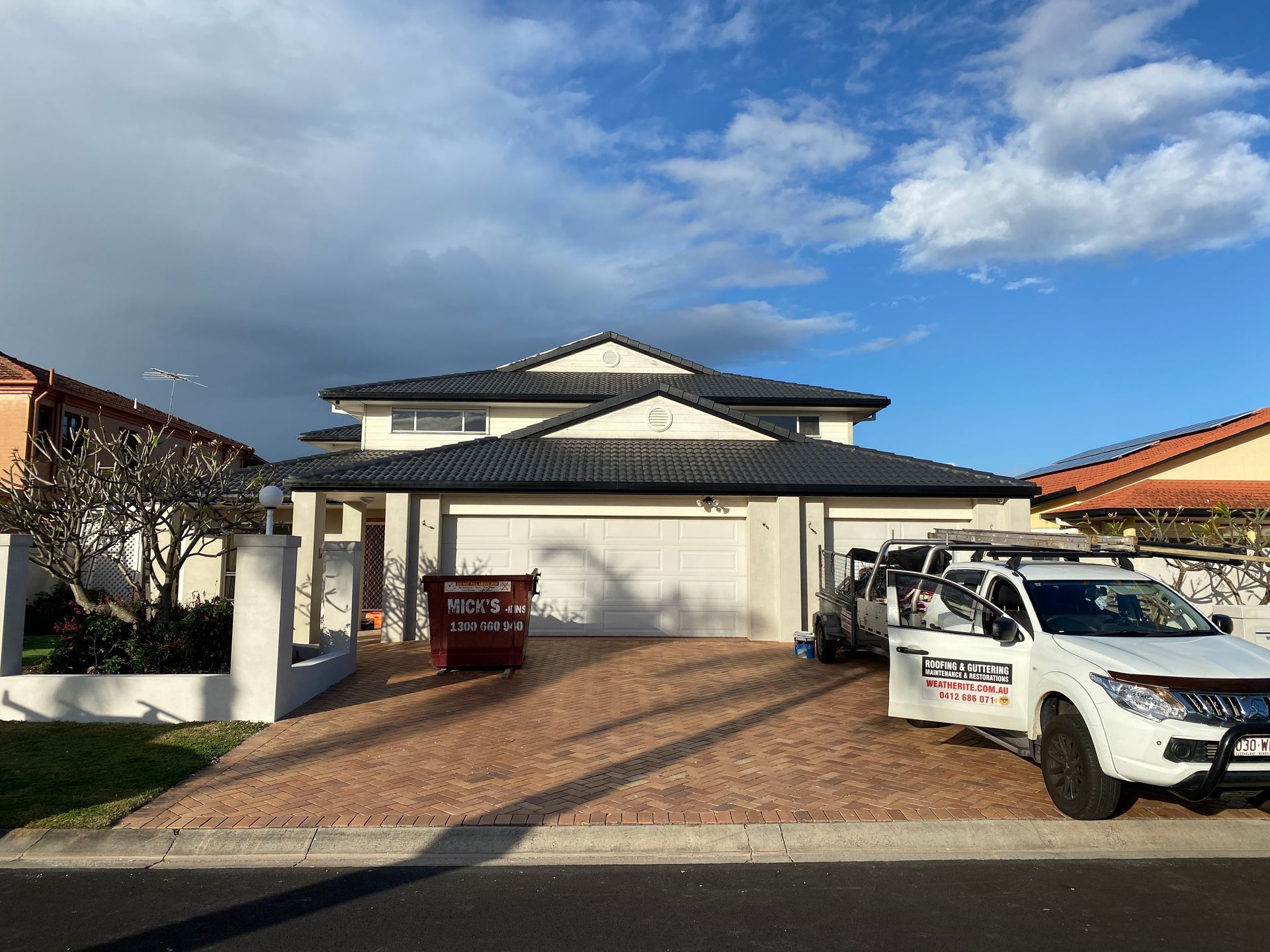 Two-story house with grey roof, white garage doors, red brick driveway, and a white utility truck.