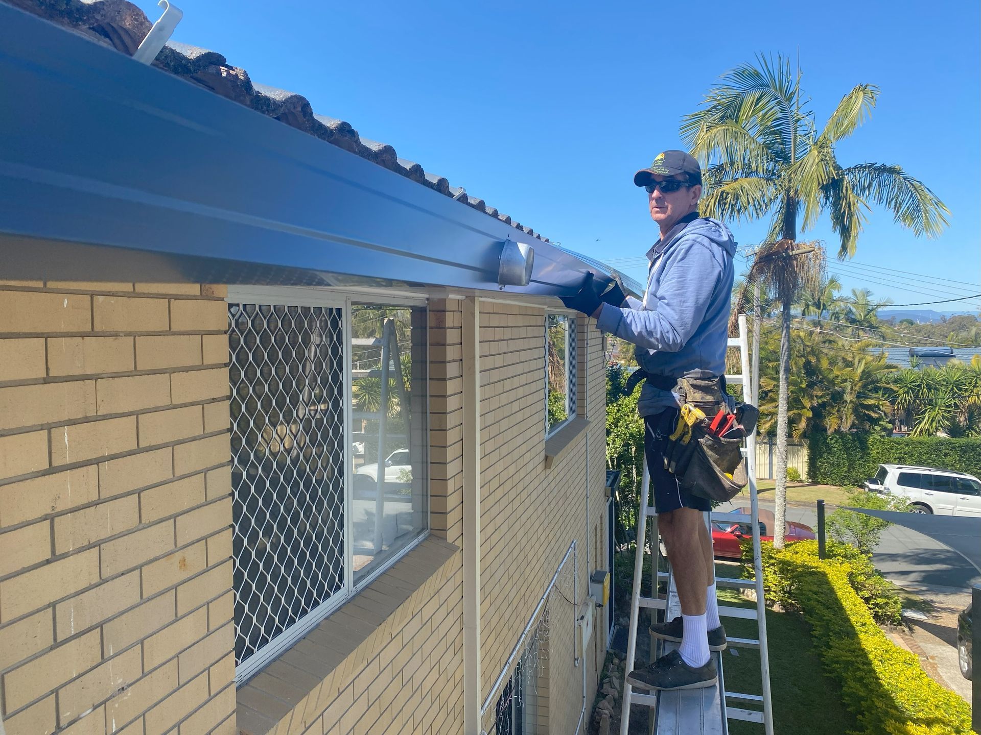 Man on ladder installing dark gutter on a brick house, sunny day.
