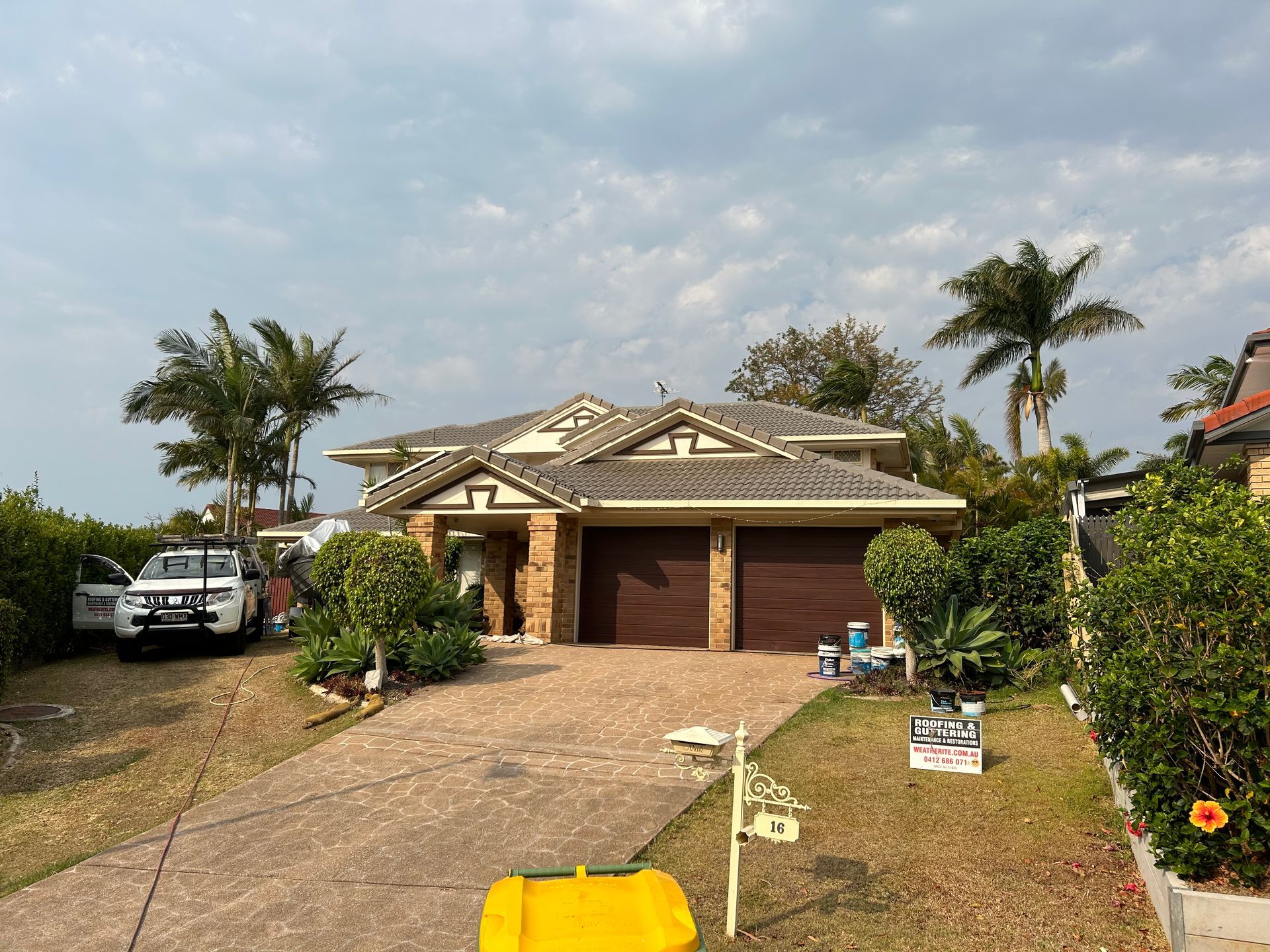 House with brown garage doors and driveway, palm trees, and a cloudy sky.