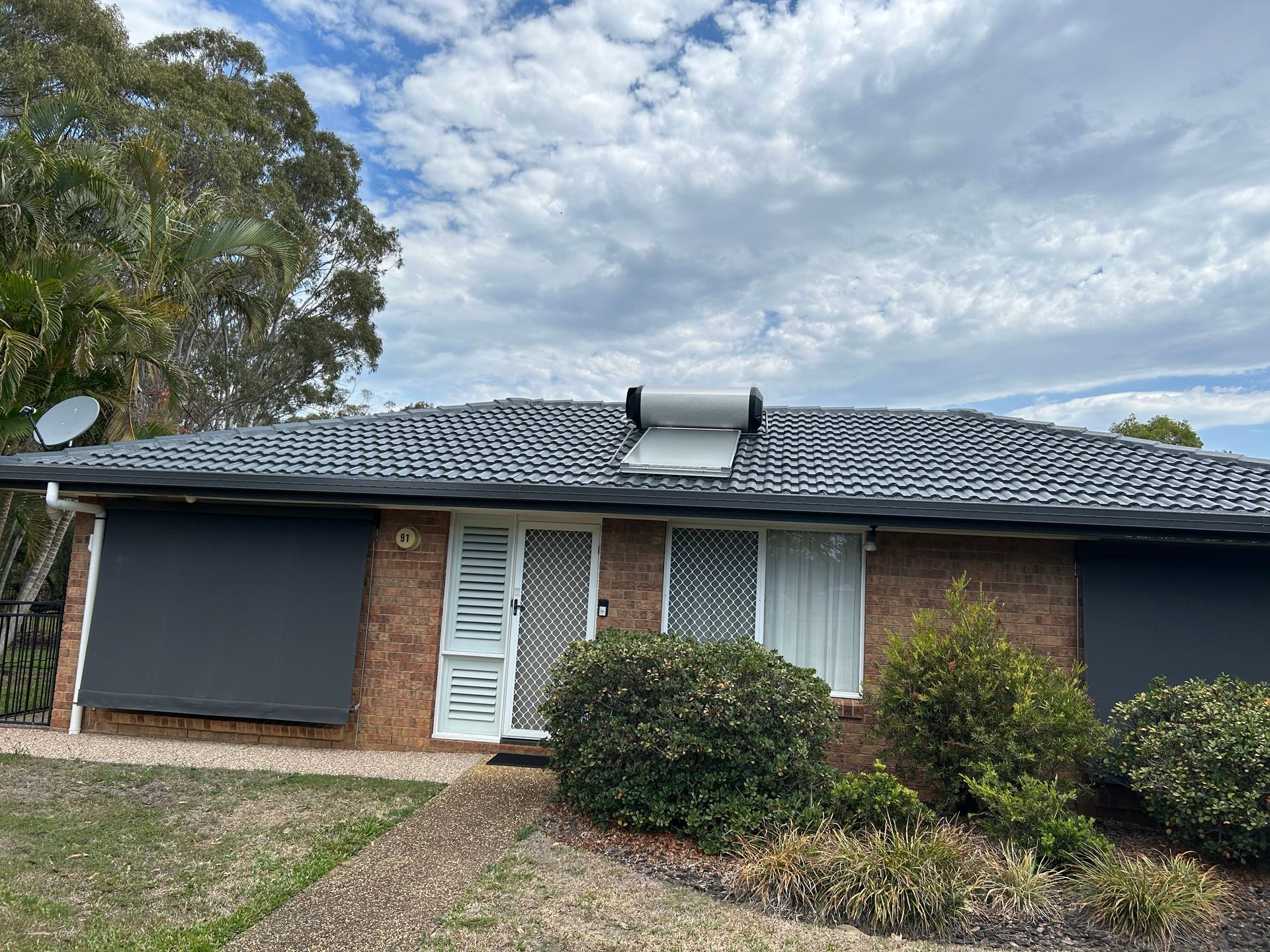 A brick house with a dark roof, solar panels, and bushes in front, under a cloudy sky.