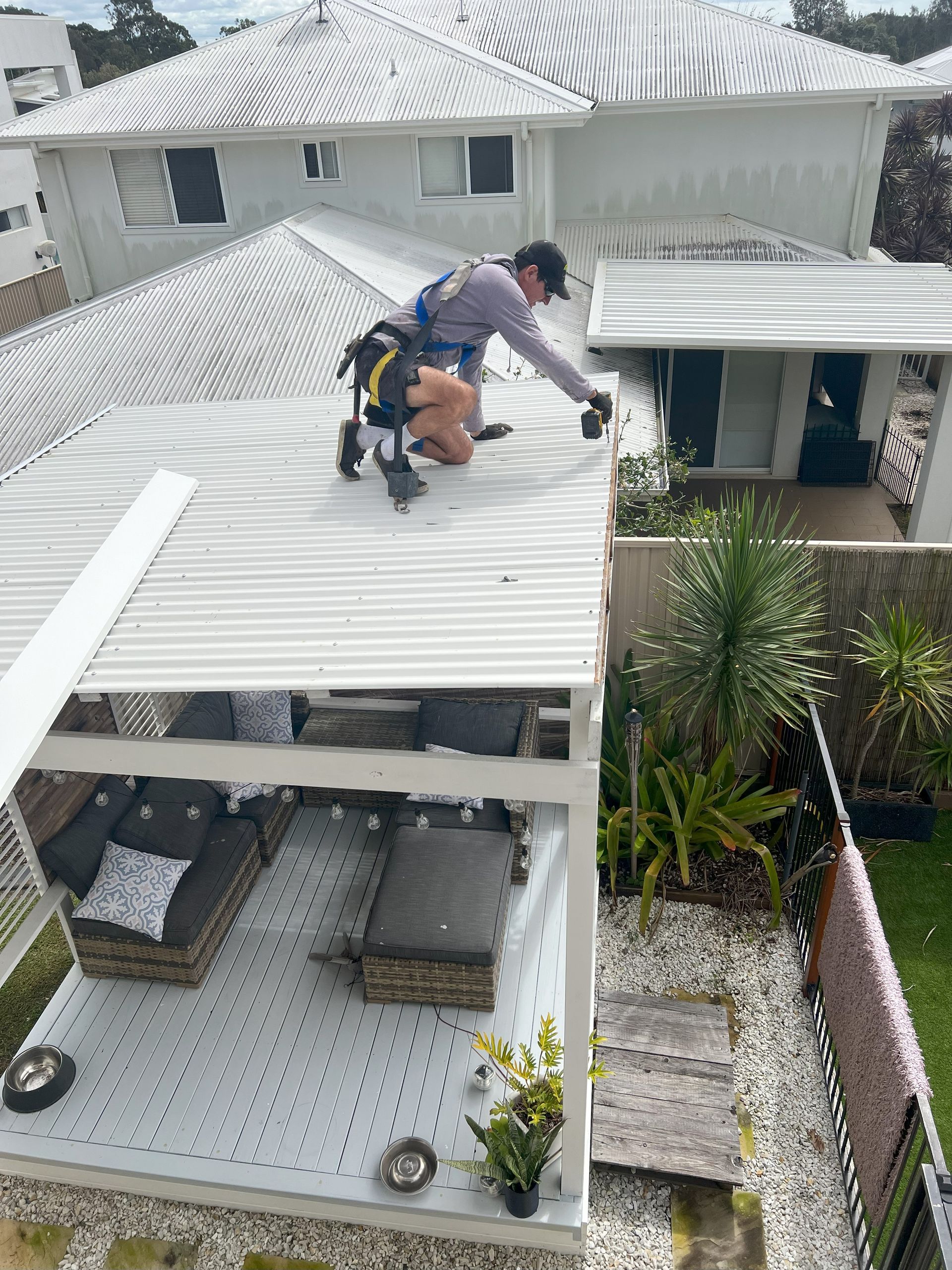 Roofer on white corrugated roof, wearing a safety harness. Patio furniture below.