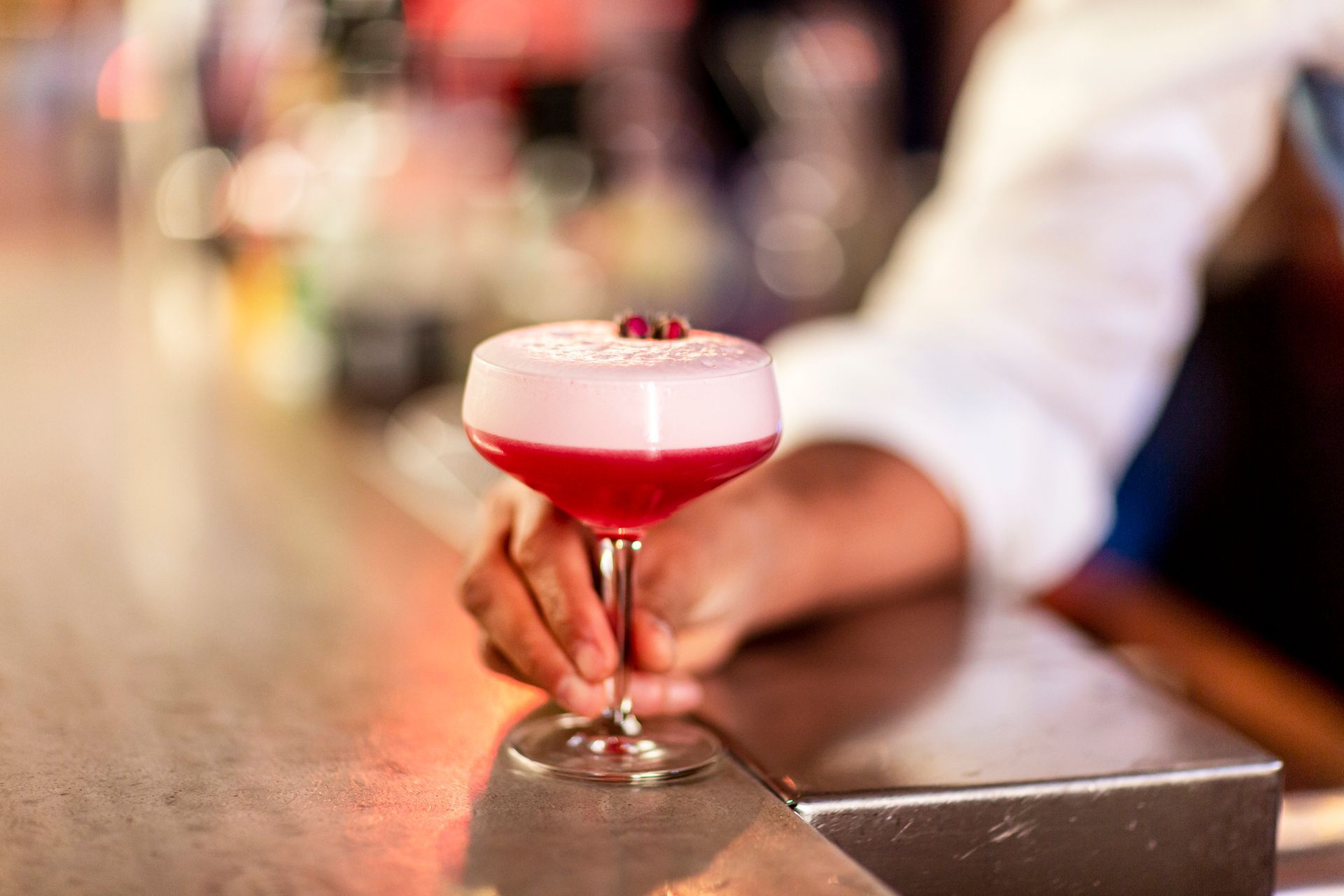 A bartender is holding a red cocktail on a bar.