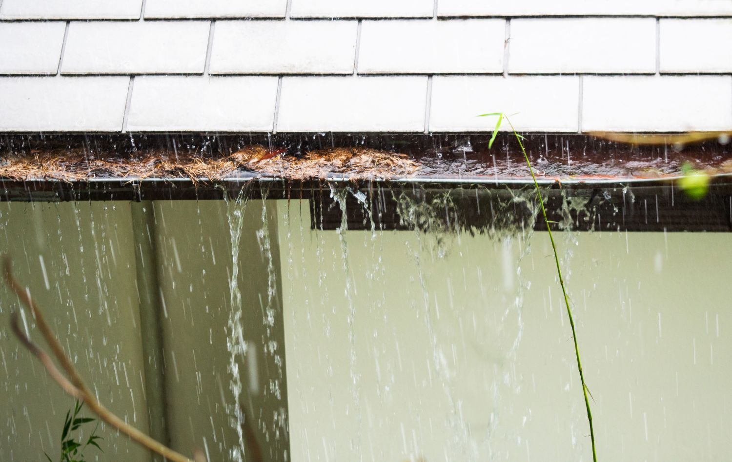 Rainwater pours from a clogged gutter. Brown debris clogs the gutter; beige wall and light tiles.