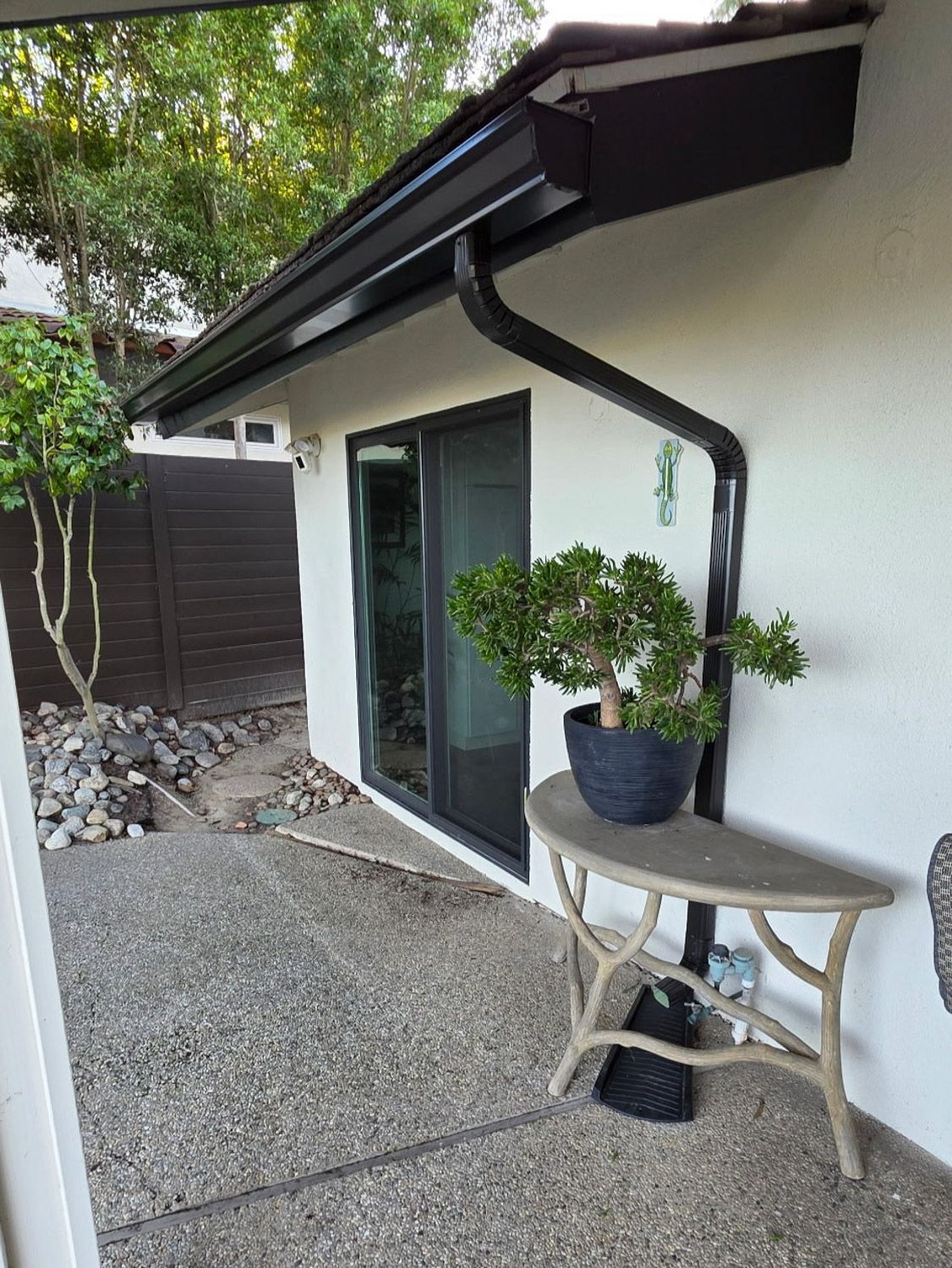 Exterior of a house with black gutters and a sliding glass door. A small tree sits on a table. Gravel walkway. New seamless gutters installed by Gutter Specialist in Reno, NV