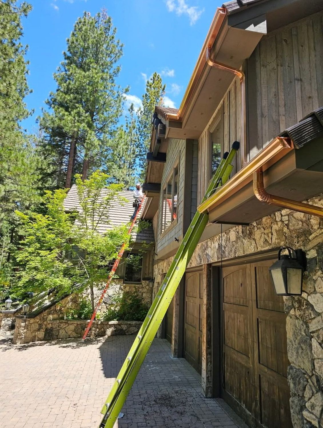 A person on a ladder cleaning a copper gutter on a stone and wood house with trees and blue sky visible. New seamless gutters installed by Gutter Specialist in Reno, NV
