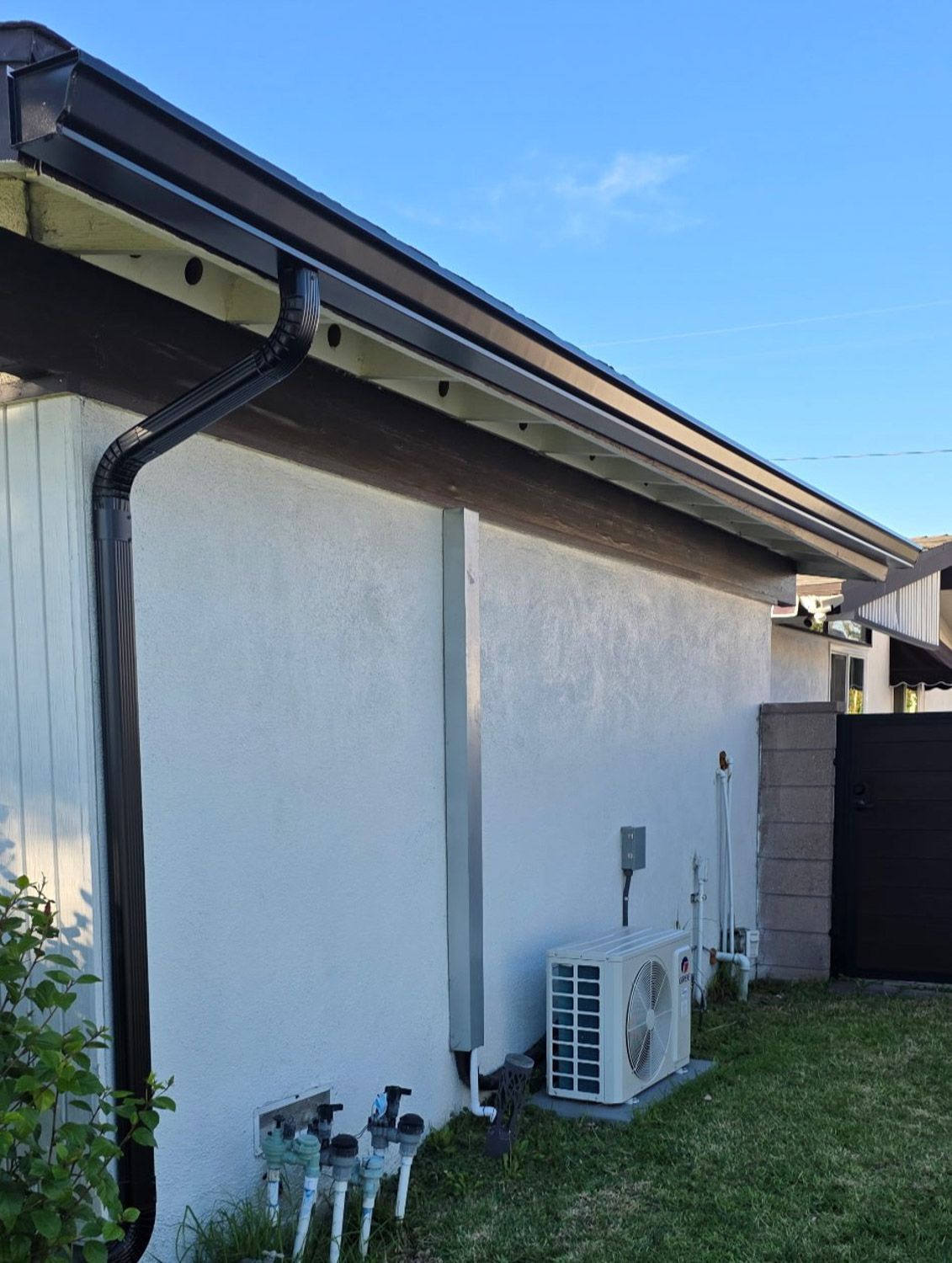 Black gutters on a white stucco building with a downspout, air conditioning unit, and a blue sky.