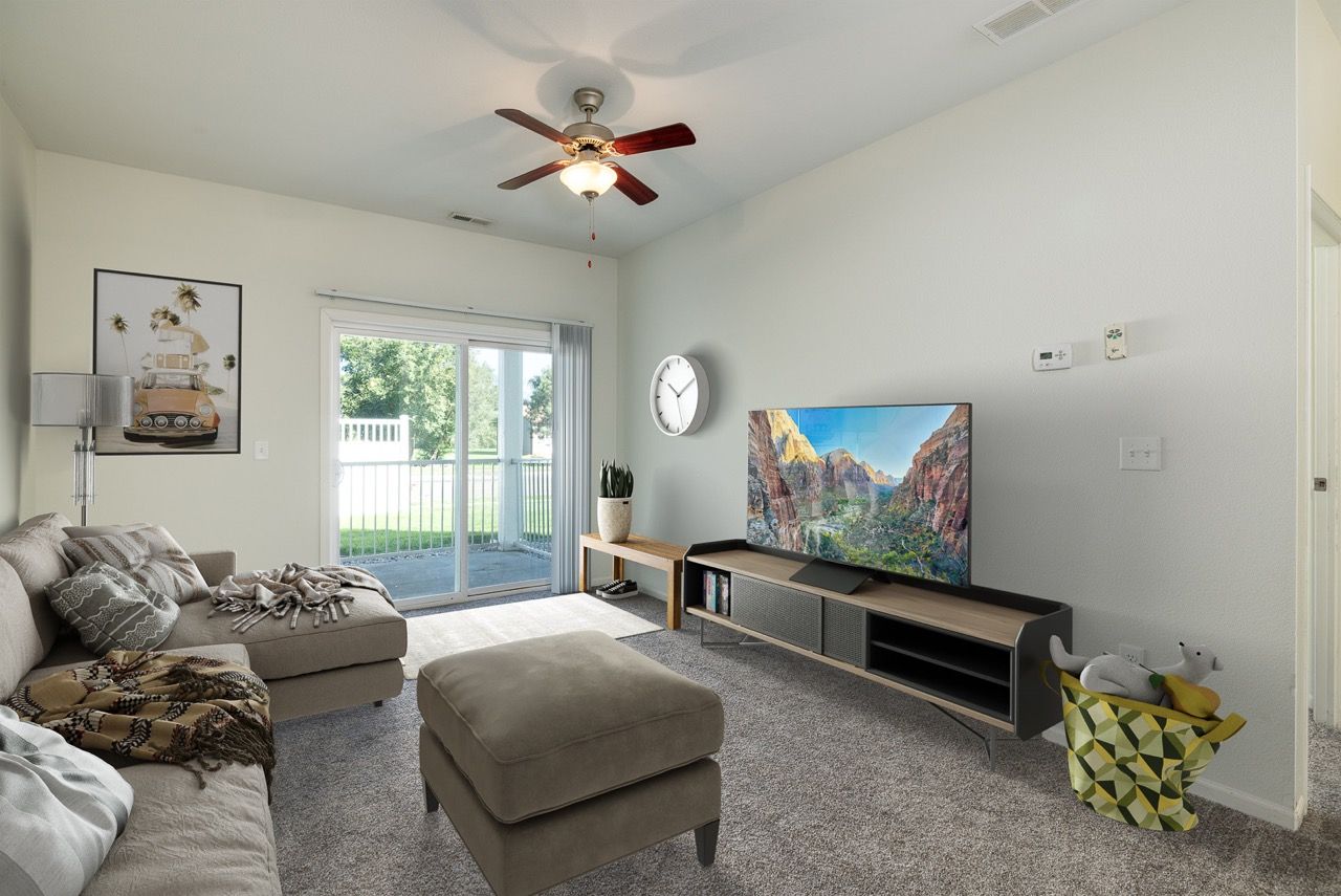 Living room in an apartment with a beige sectional sofa, ottoman, TV, sliding glass door to a patio, and ceiling fan.
