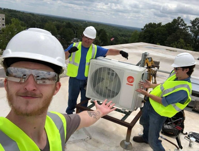 Three workers wearing hard hats and high-visibility vests pose for a selfie on a roof next to an outdoor HVAC unit.