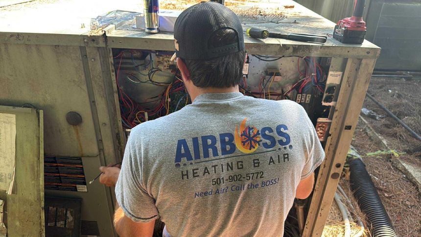 A technician in an Airboss Heating & Air t-shirt services an open HVAC unit outdoors.