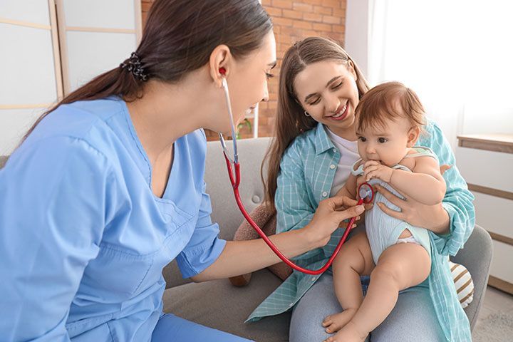 Female pediatrician with stethoscope listening to little baby and mother on sofa at home 
