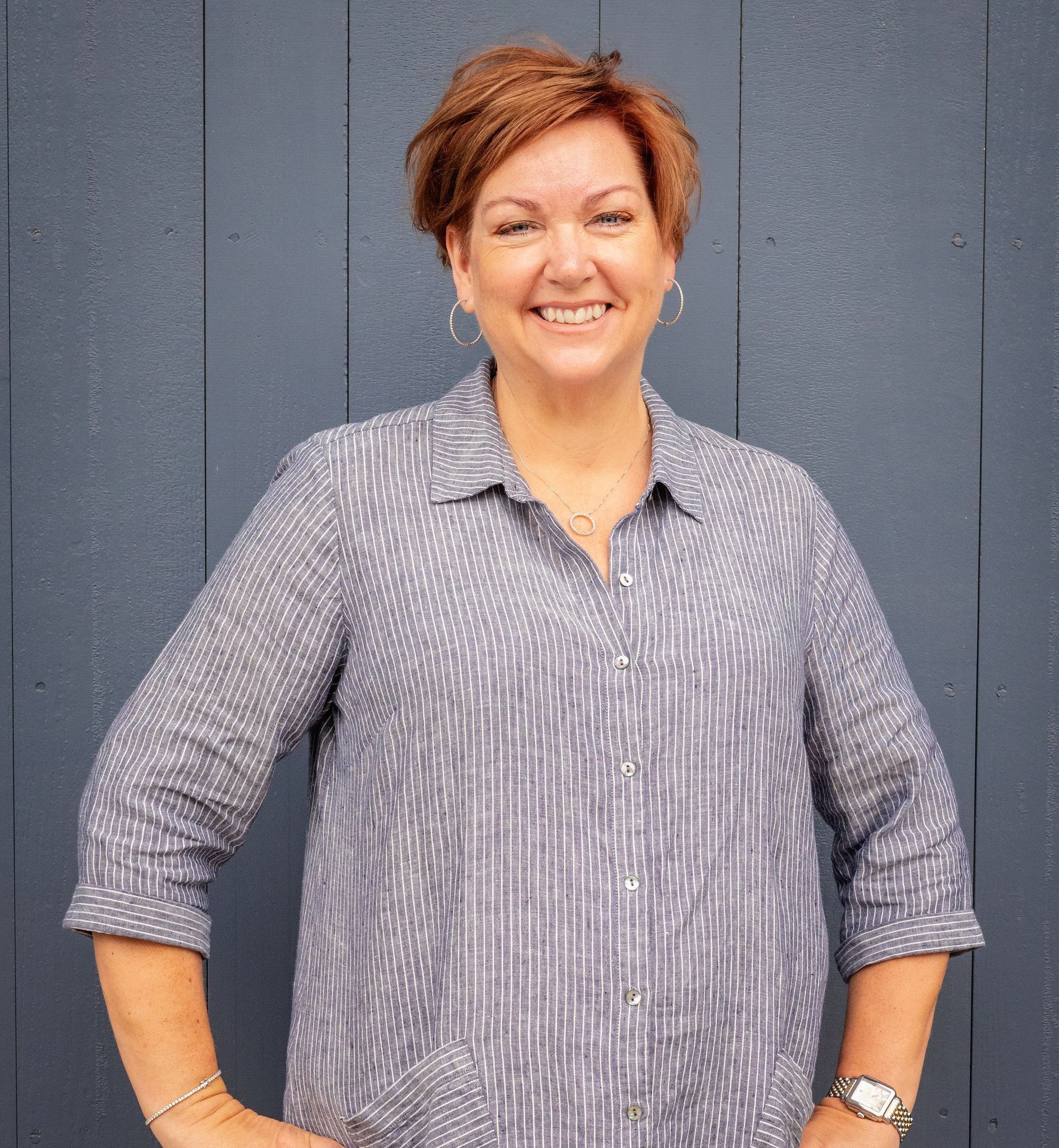 A woman in a striped shirt is standing in front of a blue wall with her hands in her pockets.