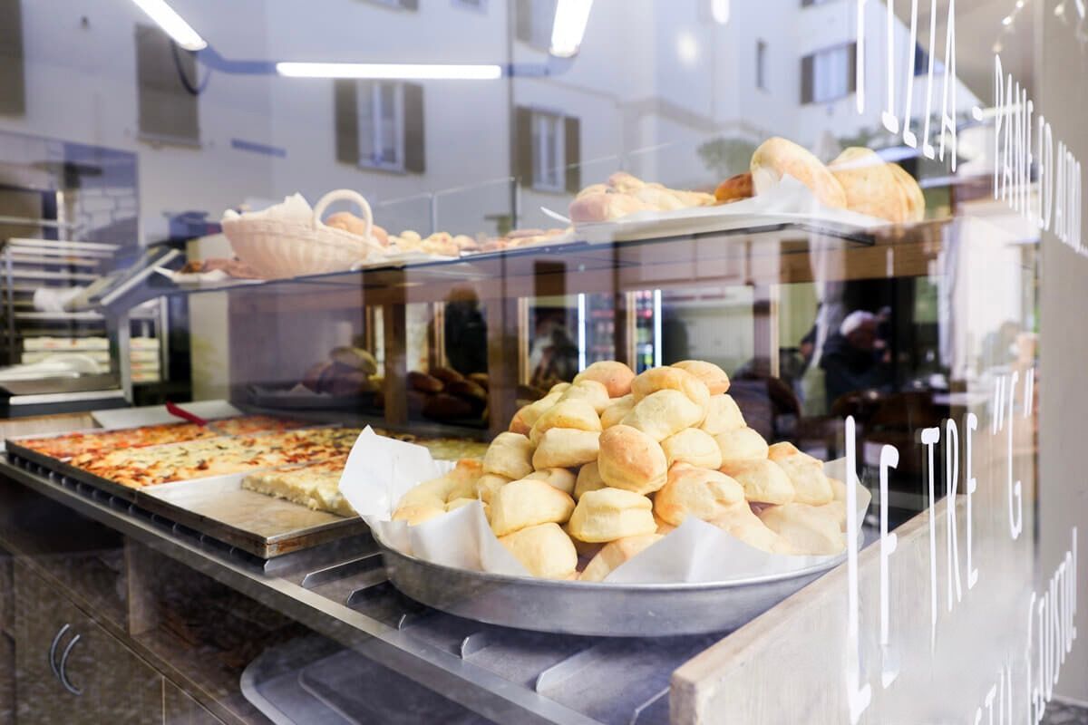 Esposizione di prodotti da forno: pasticcini, pizza e pane sugli scaffali dietro una vetrina che riflette gli edifici.