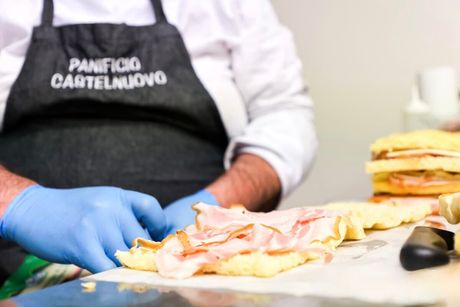 Cuoco con guanti blu che prepara un panino, aggiungendo fette di carne. Sul grembiule nero c'è la scritta 