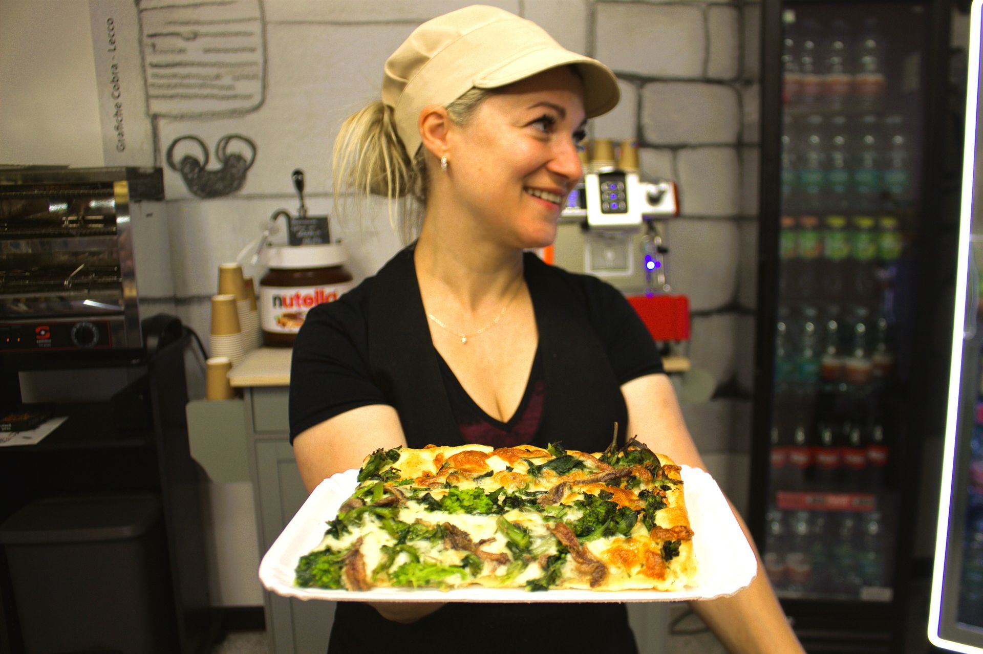 Donna che regge un vassoio con una quiche quadrata. Indossa un berretto e sorride in un bar.