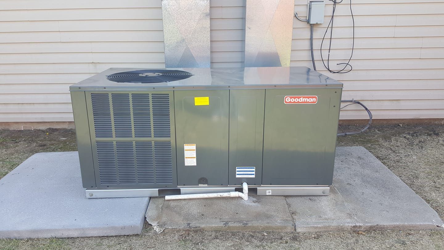 A Goodman air conditioning unit on a concrete pad, next to a building. Gray metal, with vents and a fan on top.