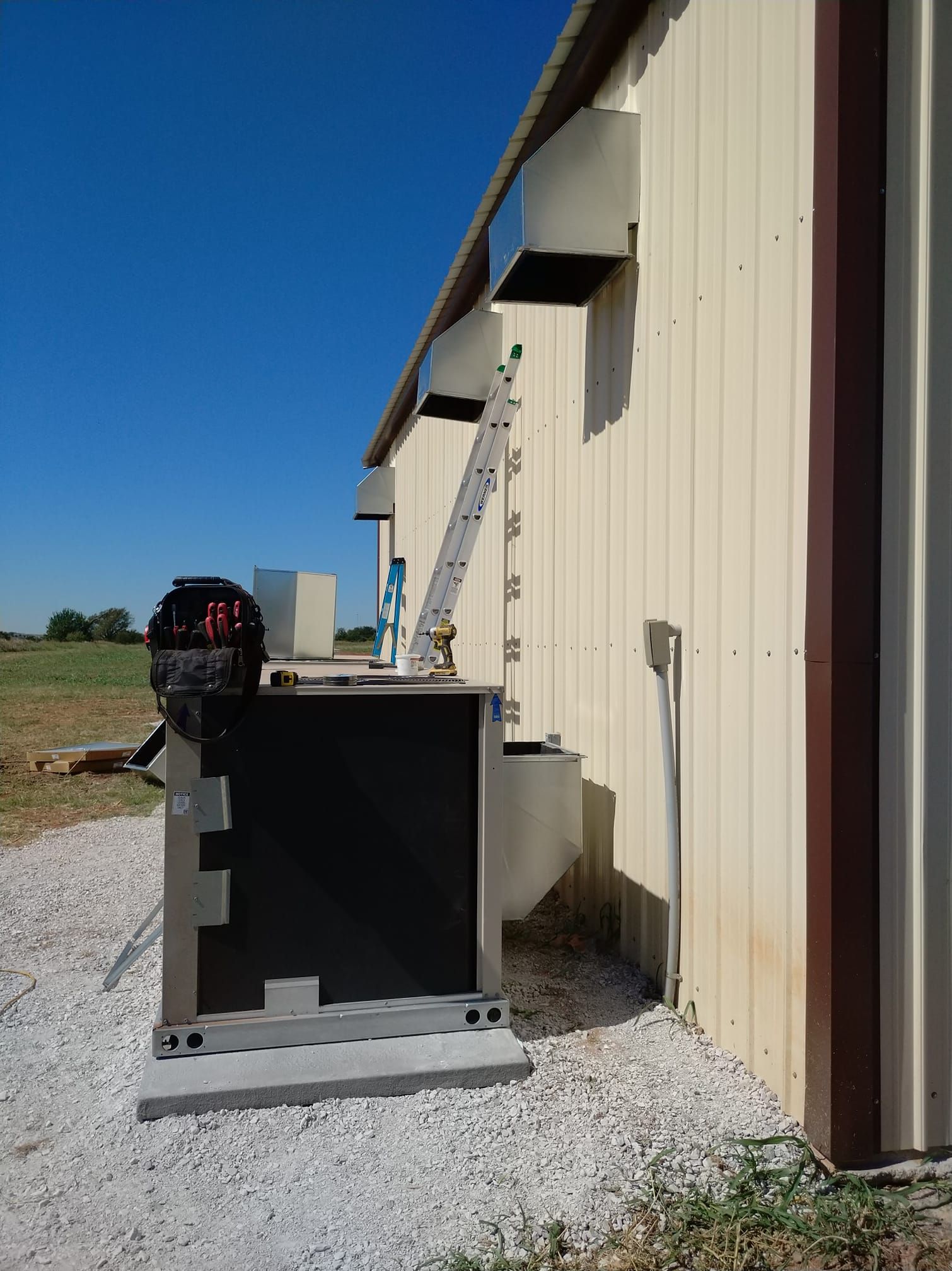 Metal building exterior with a dark-colored utility box, vents, and a ladder; gravel ground and a blue sky.