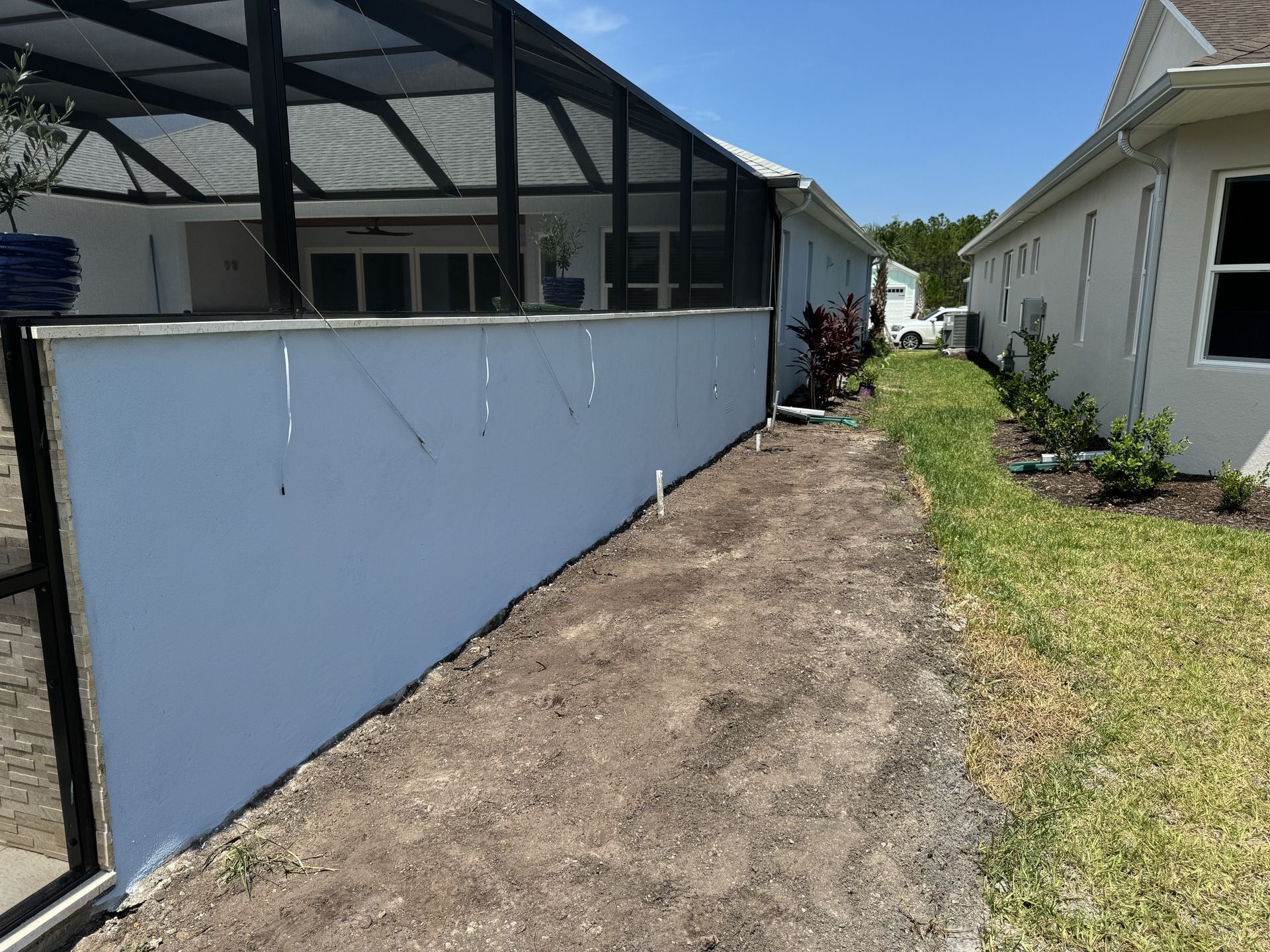 Blue stucco wall along a path, adjacent to a screened enclosure and a white house, sunny day.