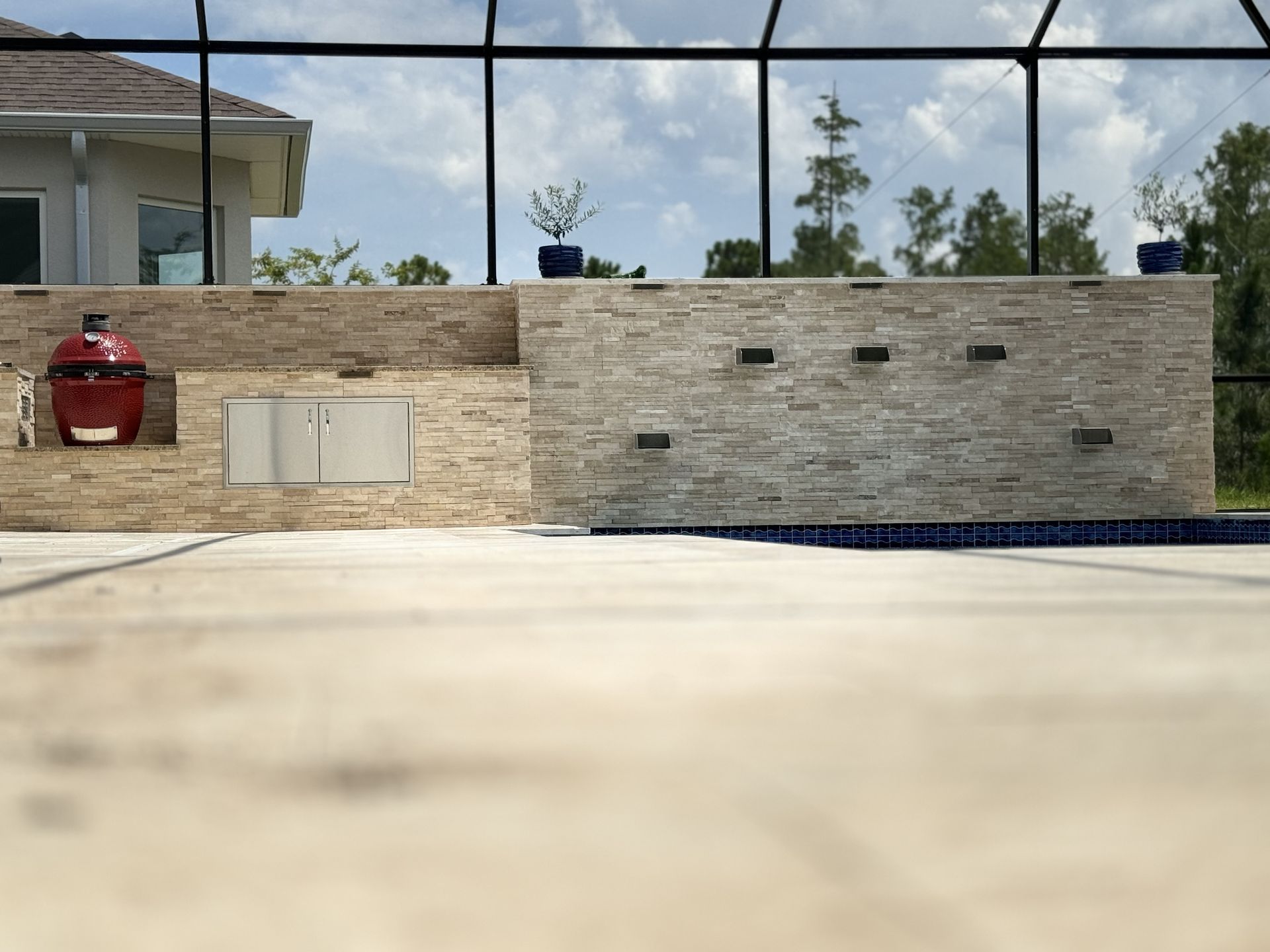 Outdoor kitchen with a red grill, built-in cabinets, and pool in a sunny setting.