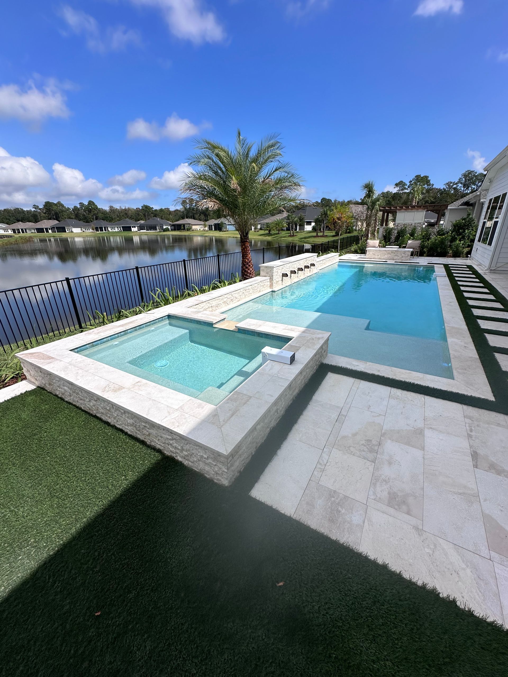 Pool with jacuzzi, overlooking a lake. White stone patio, artificial turf, sunny day.