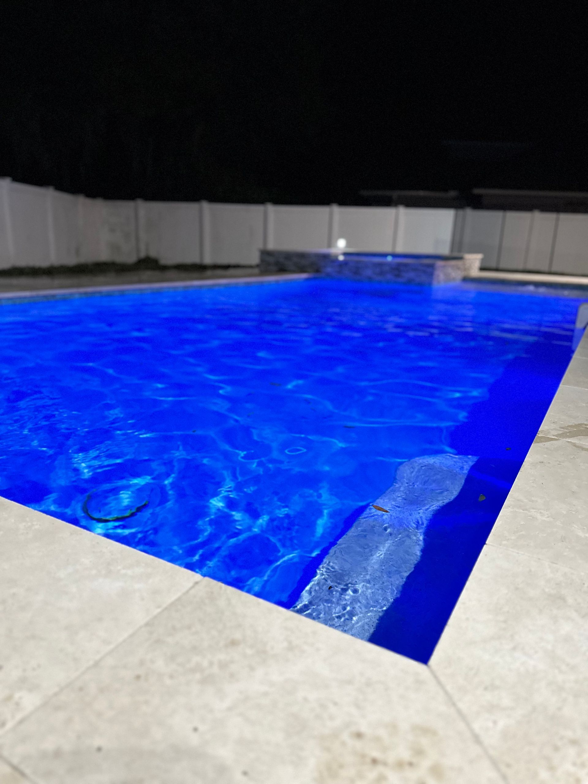 Blue-lit swimming pool at night, surrounded by light-colored tiles and a white fence.