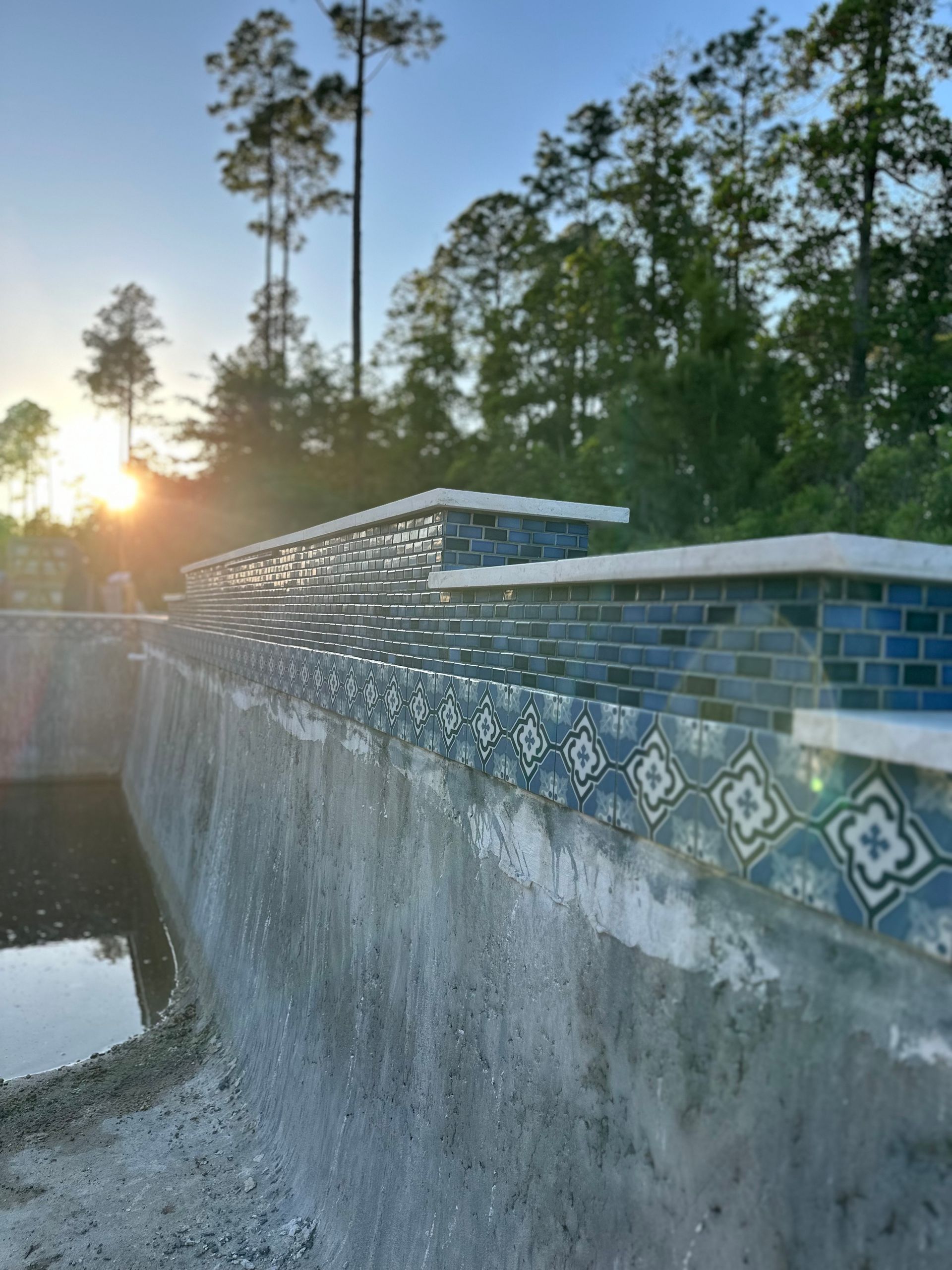 Partially-built pool with blue-tiled edges, concrete walls, and a forest backdrop at sunset.