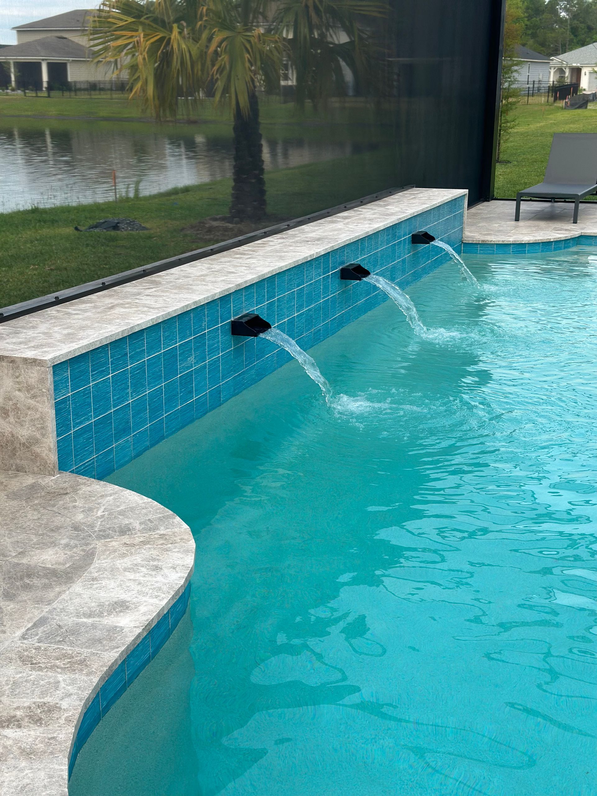 Pool with blue tile wall, fountains, and clear water; green lawn, lake, and palm tree in background.