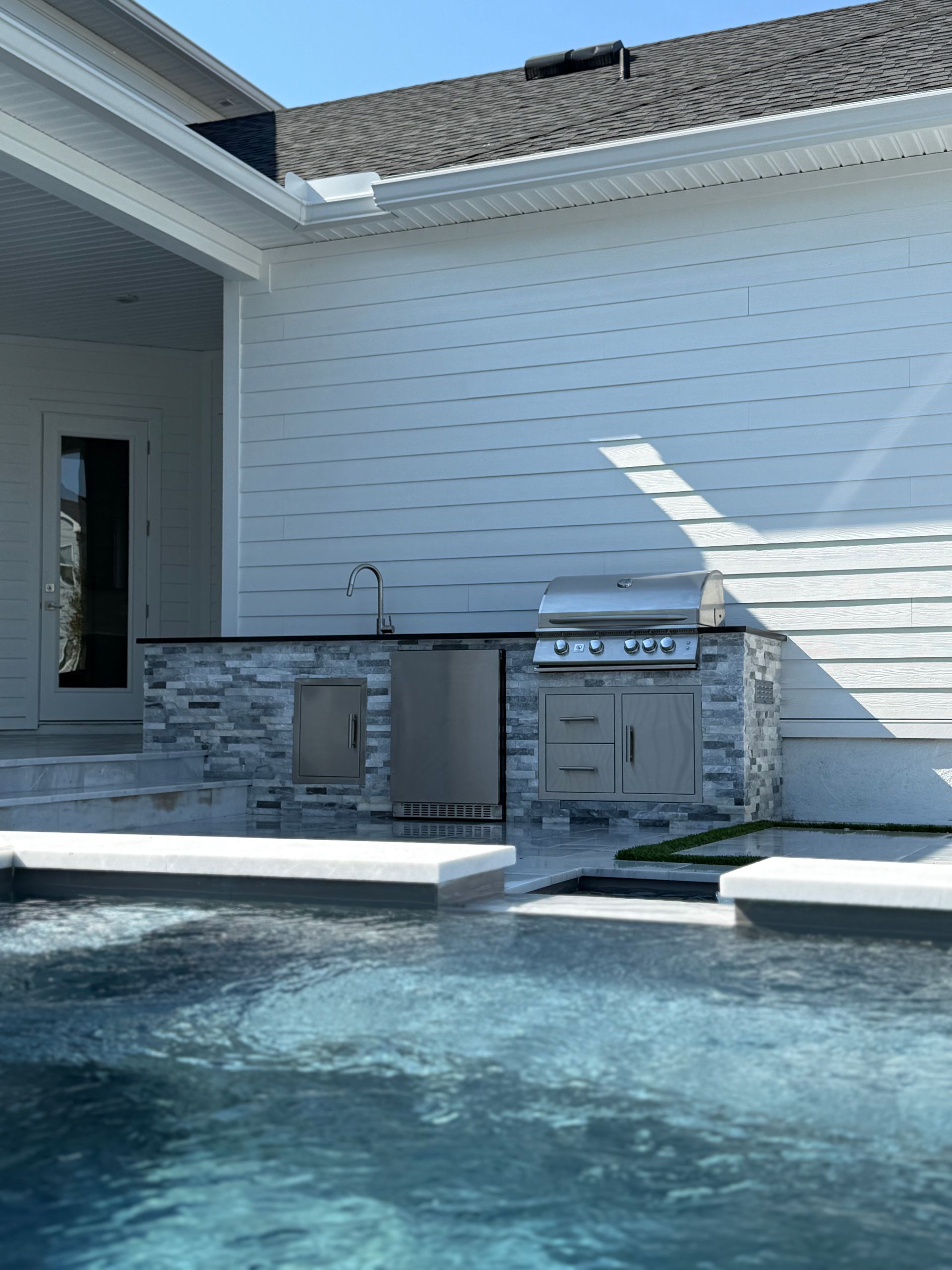 Outdoor kitchen with grill and sink next to a pool. Stone countertop and white siding, sunny day.