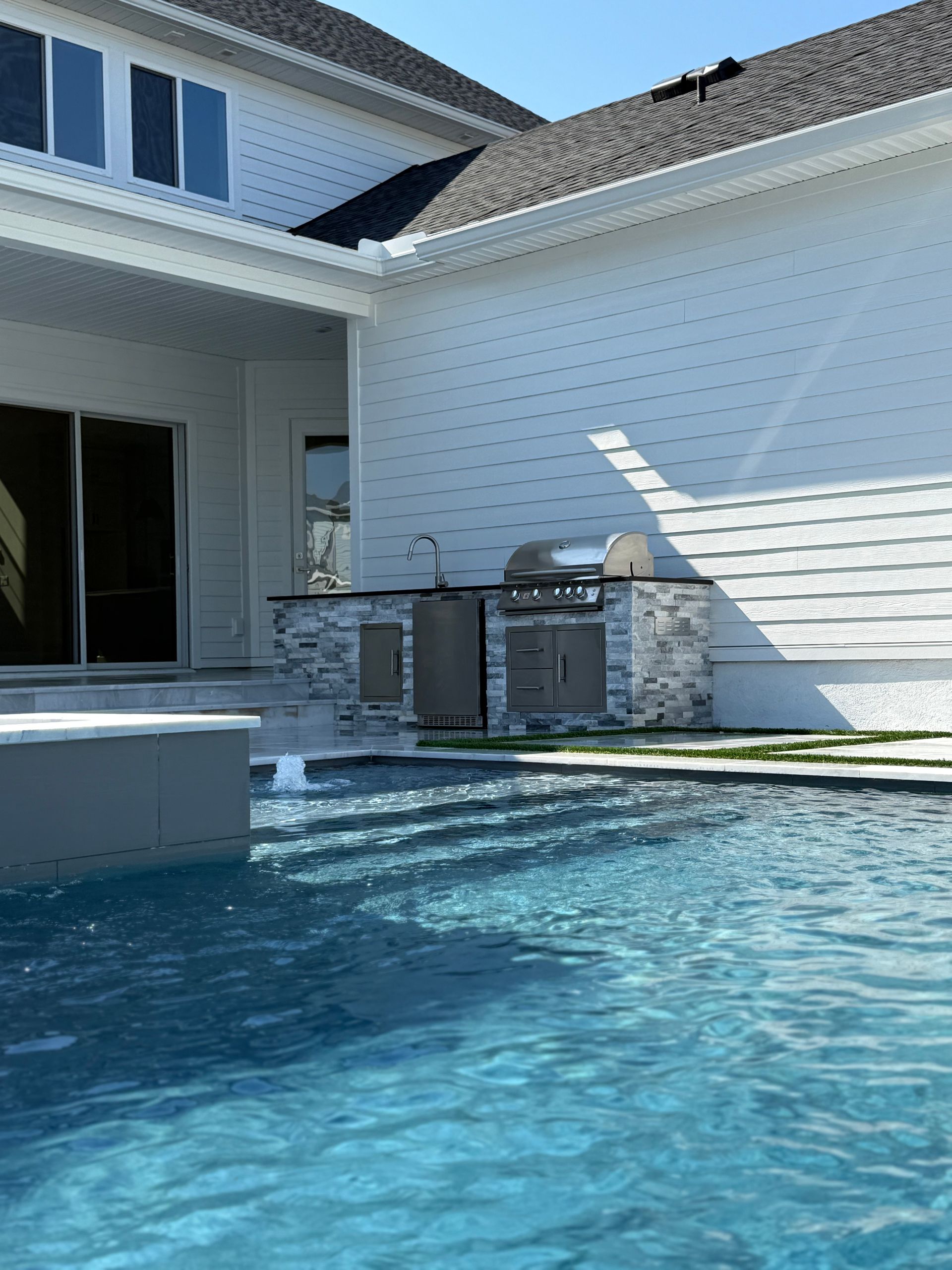 Outdoor kitchen next to a pool, with a white house and blue water. Sunny day.