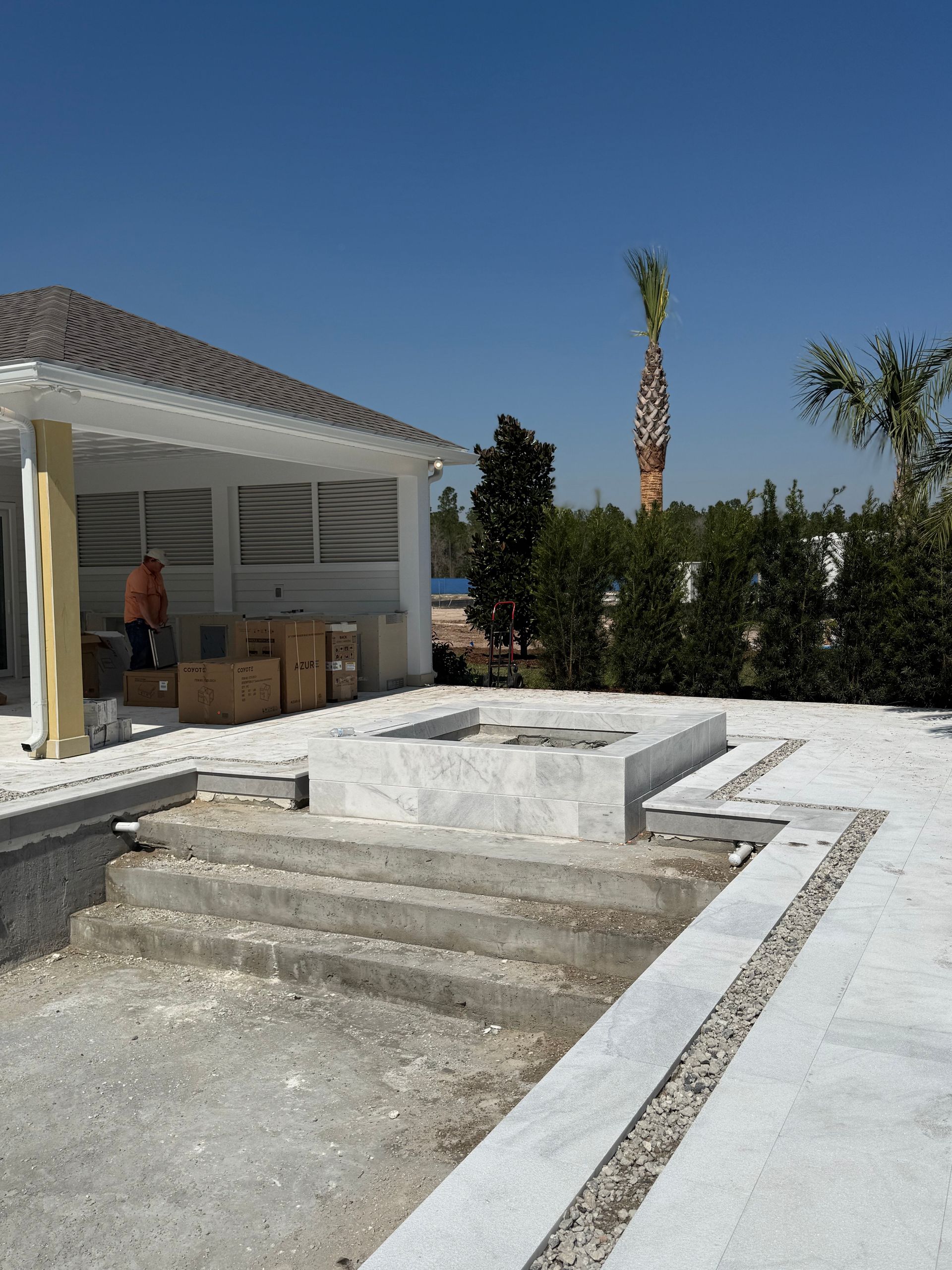 Pool under construction, featuring concrete steps and a patio area, with a covered structure and palm trees in the background.