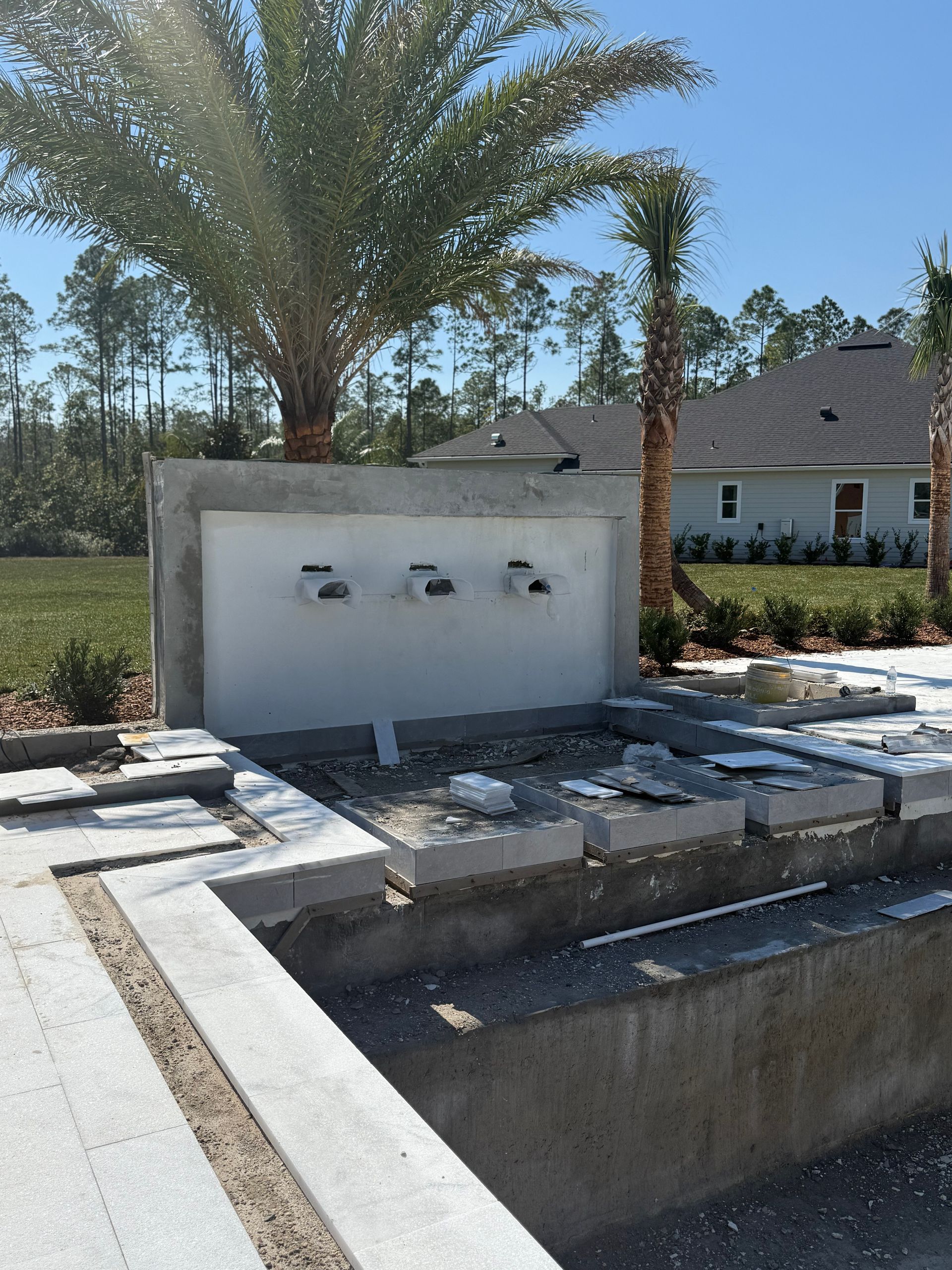 Pool construction: concrete water feature with three openings, stone border, palm trees, and blue sky.