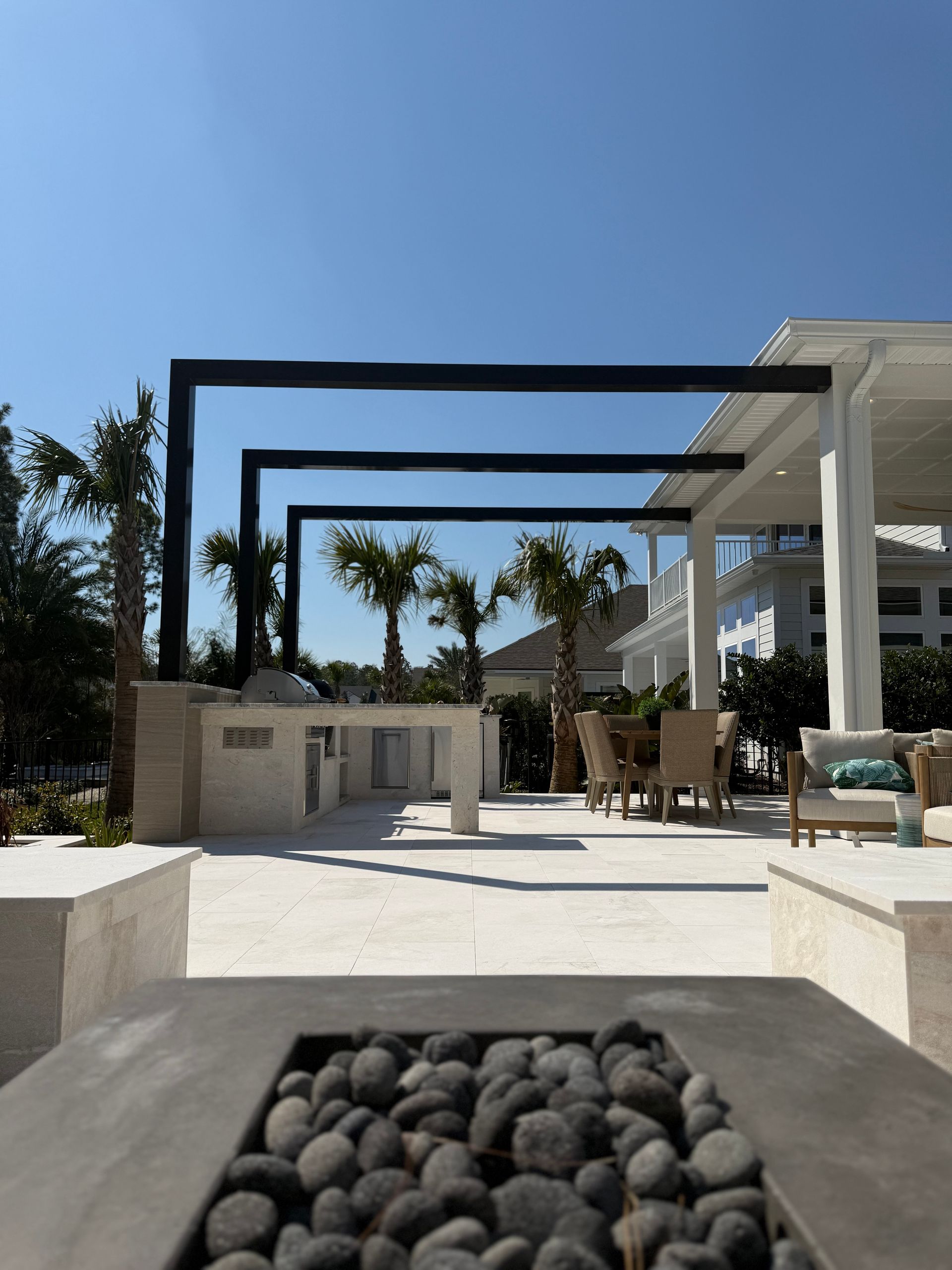Outdoor patio with a pergola, grill, seating, and a fire pit under a blue sky.