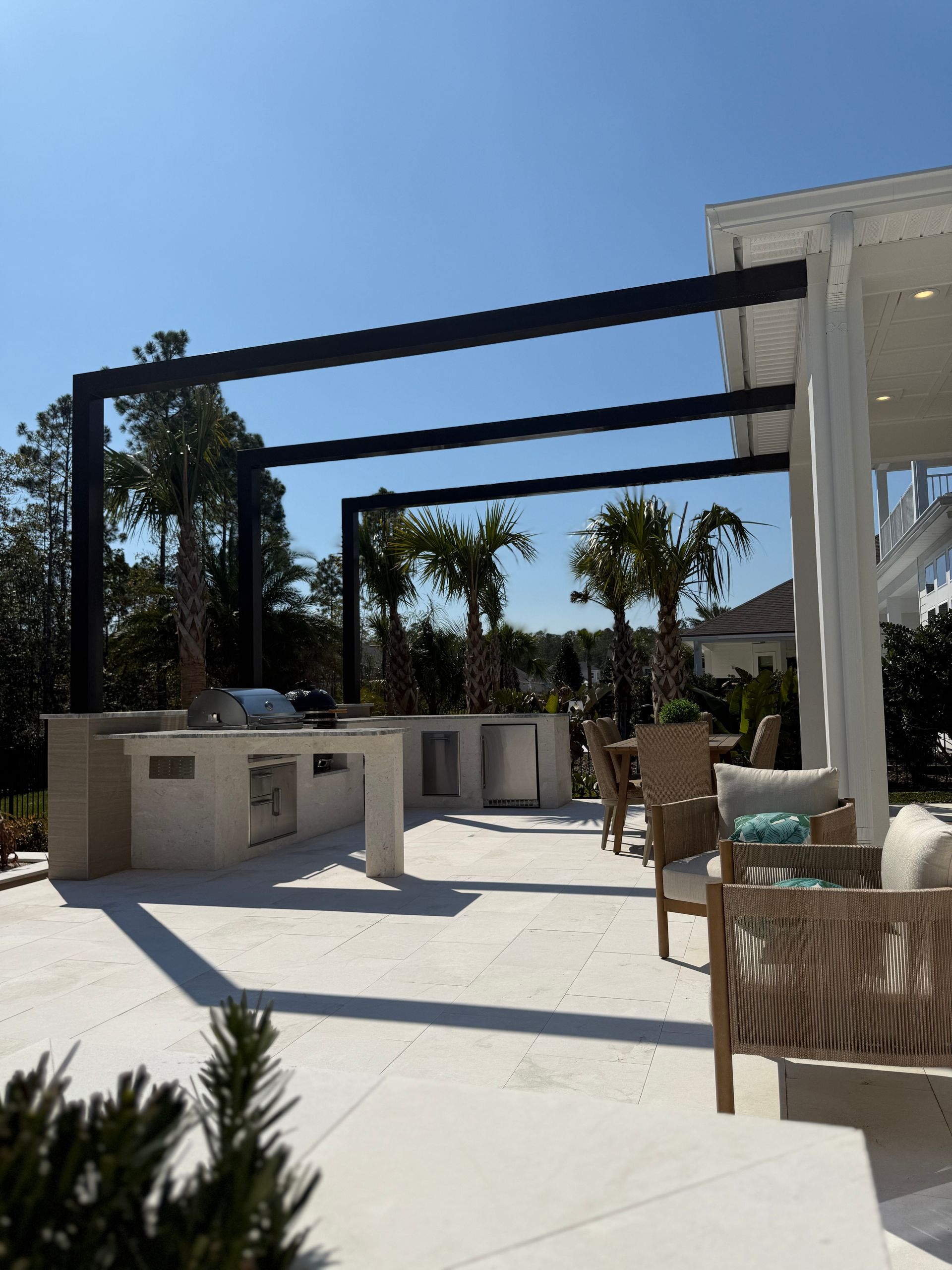 Outdoor kitchen with grill, sink, seating under black pergola, sunny day.