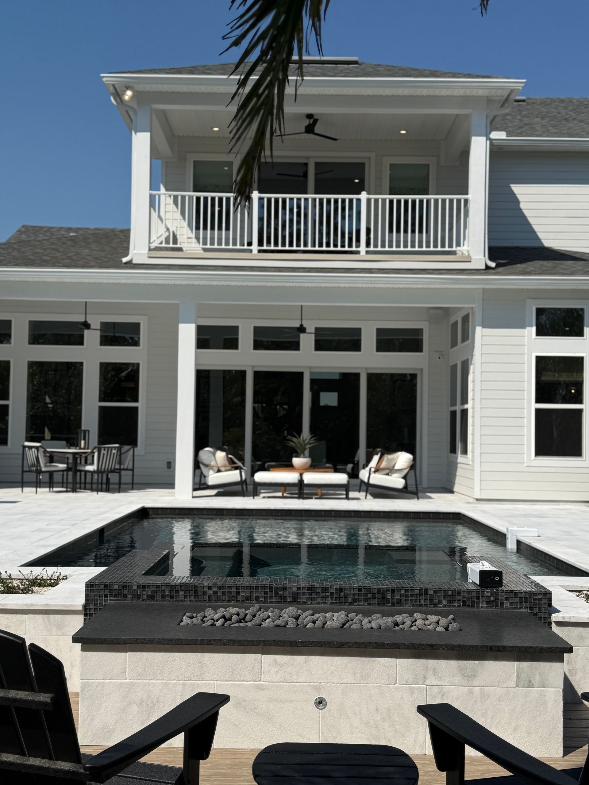 White house with pool and balcony. Black chairs face a built-in fire pit. Sunny, outdoor setting.