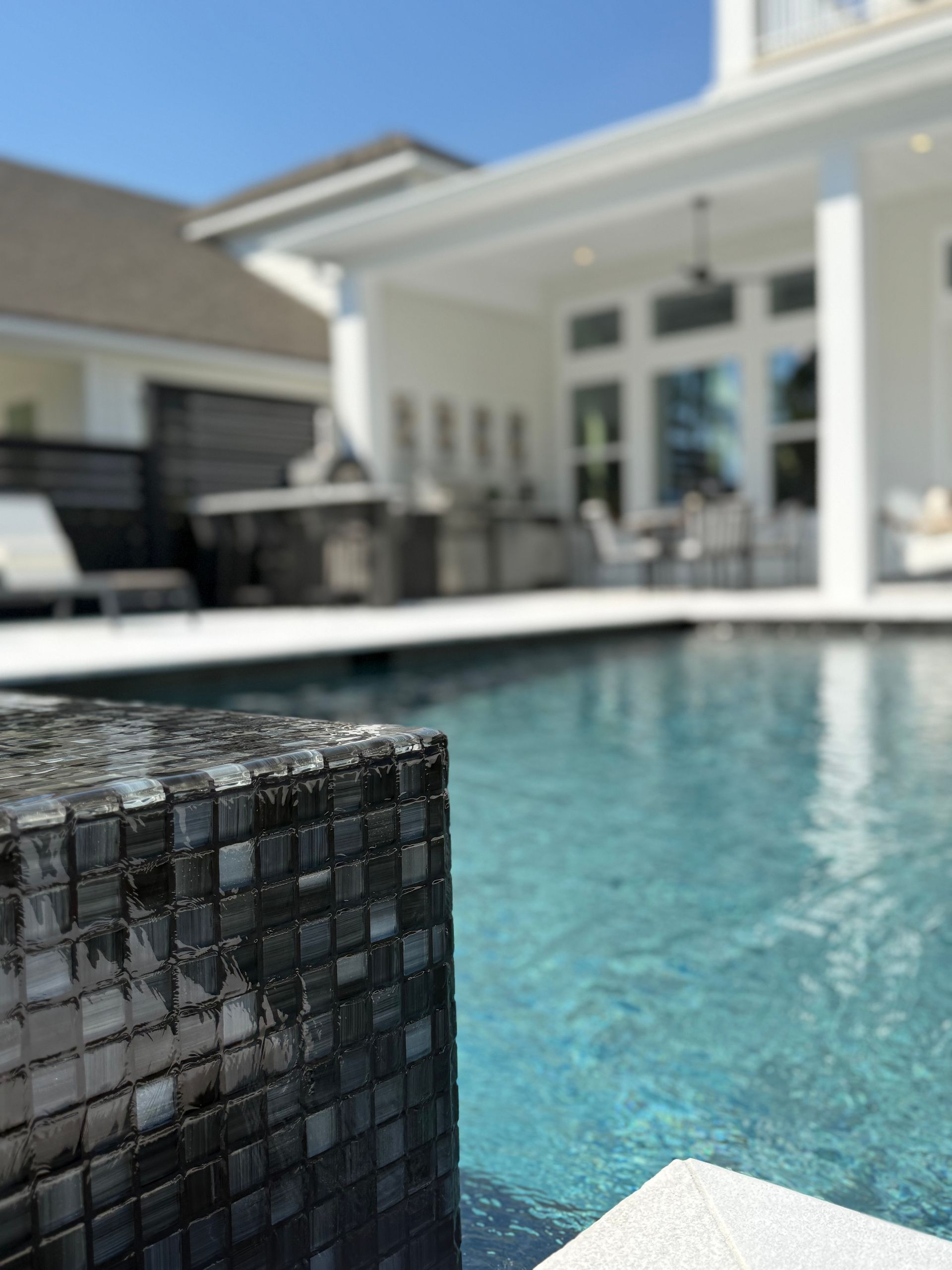Pool with dark tile border, blurred background of white house, outdoor patio.