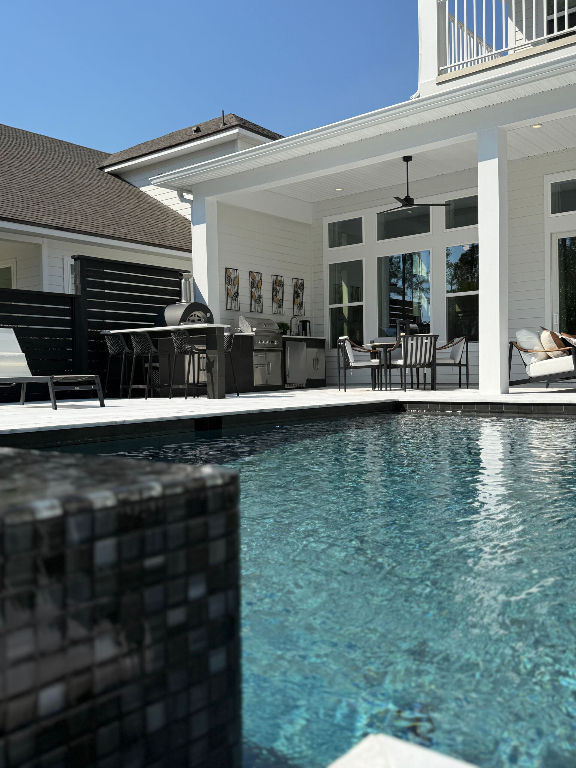 Poolside patio with grill, dining area, and house. Bright blue sky over clear water.