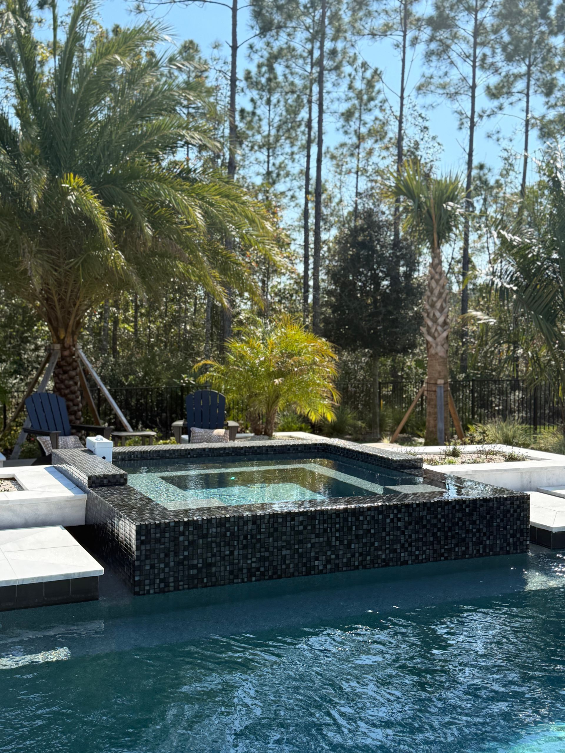 Pool with spa, surrounded by patio, trees. Dark tile, turquoise water, sunny day.