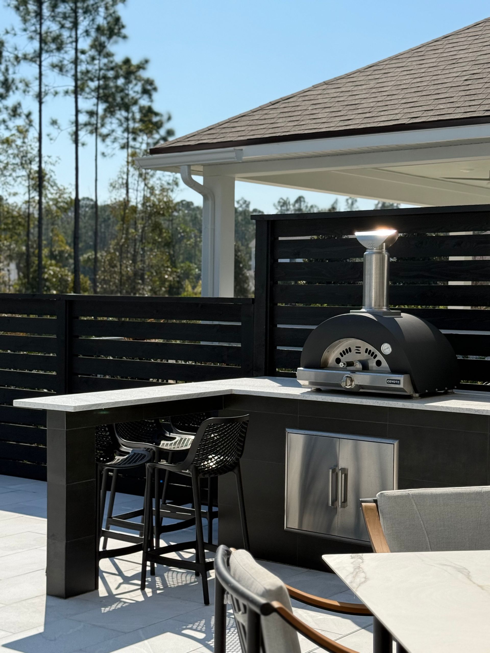 Outdoor kitchen with pizza oven, seating, and black horizontal fence. Sunny day.