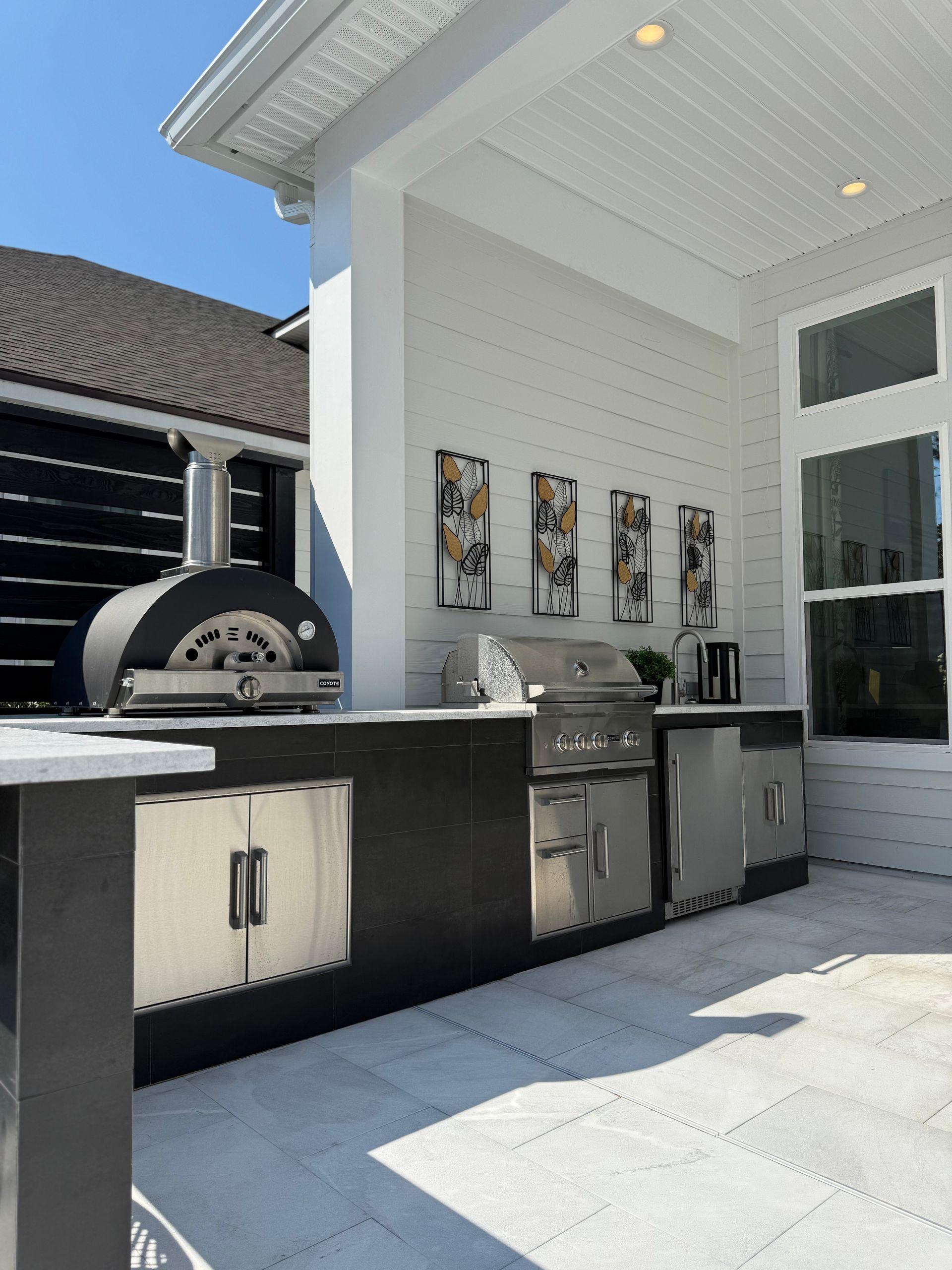 Outdoor kitchen with a pizza oven, grill, and refrigerator, set against a white wall; sunny day.