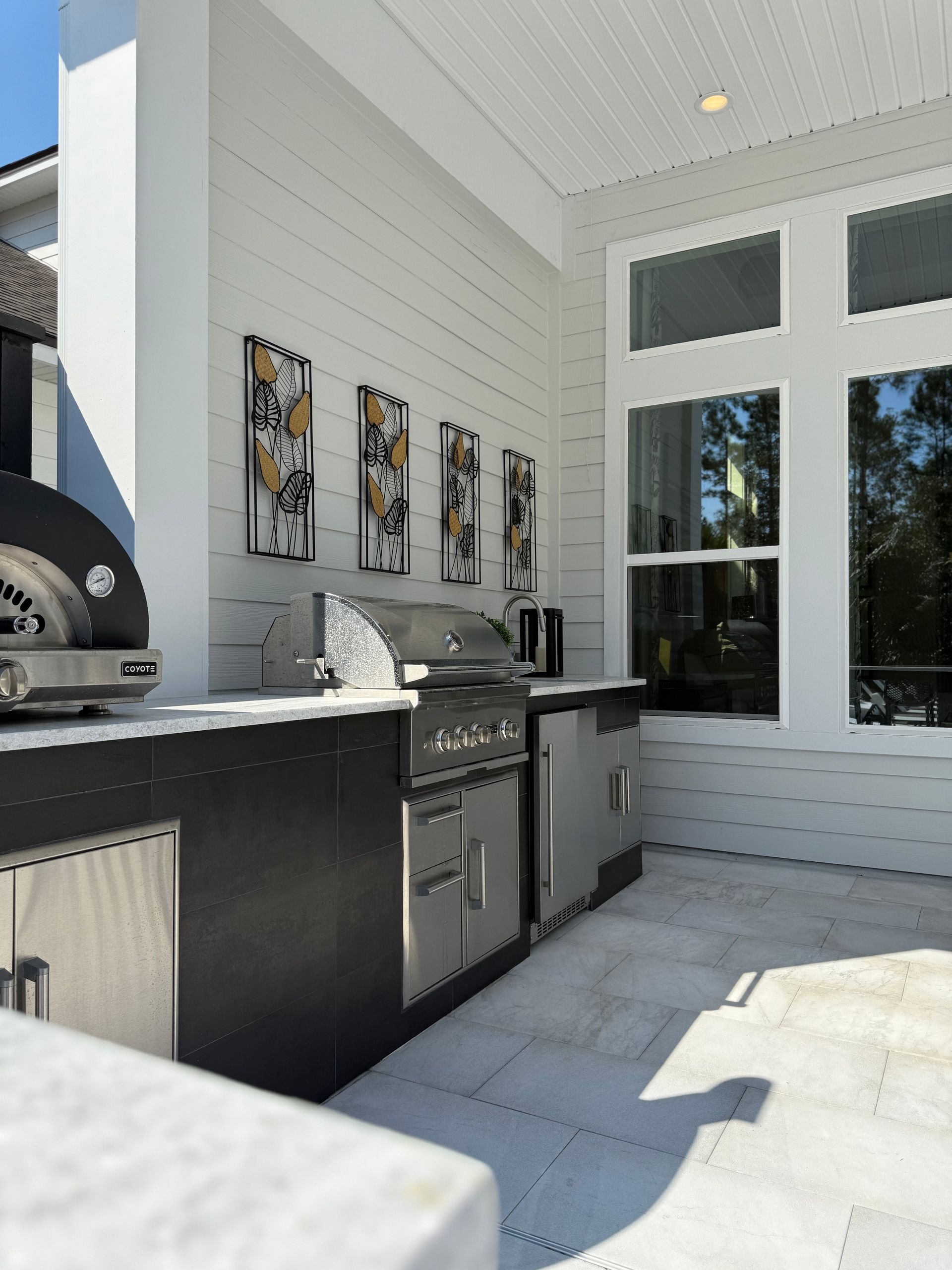 Outdoor kitchen with stainless steel appliances and pizza oven. White walls, black cabinets, art.
