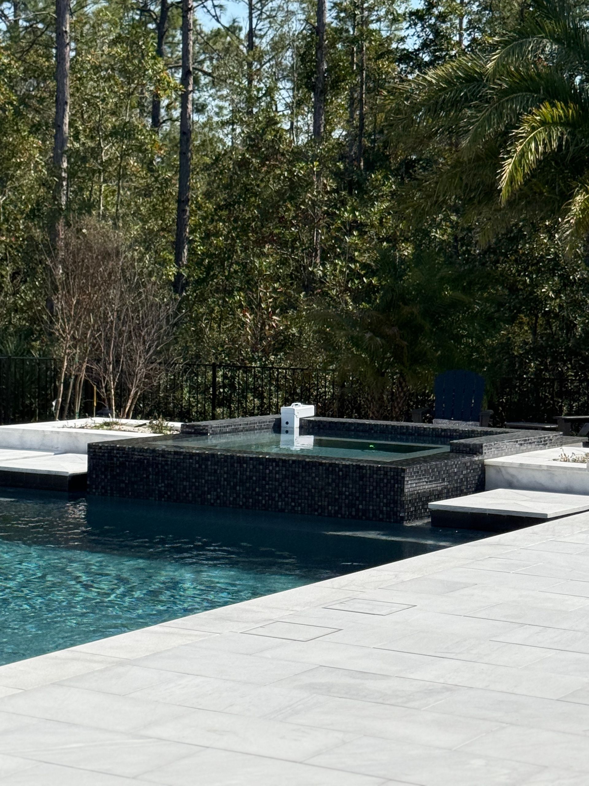Pool with dark tile spa, white deck, and lush green trees in background.
