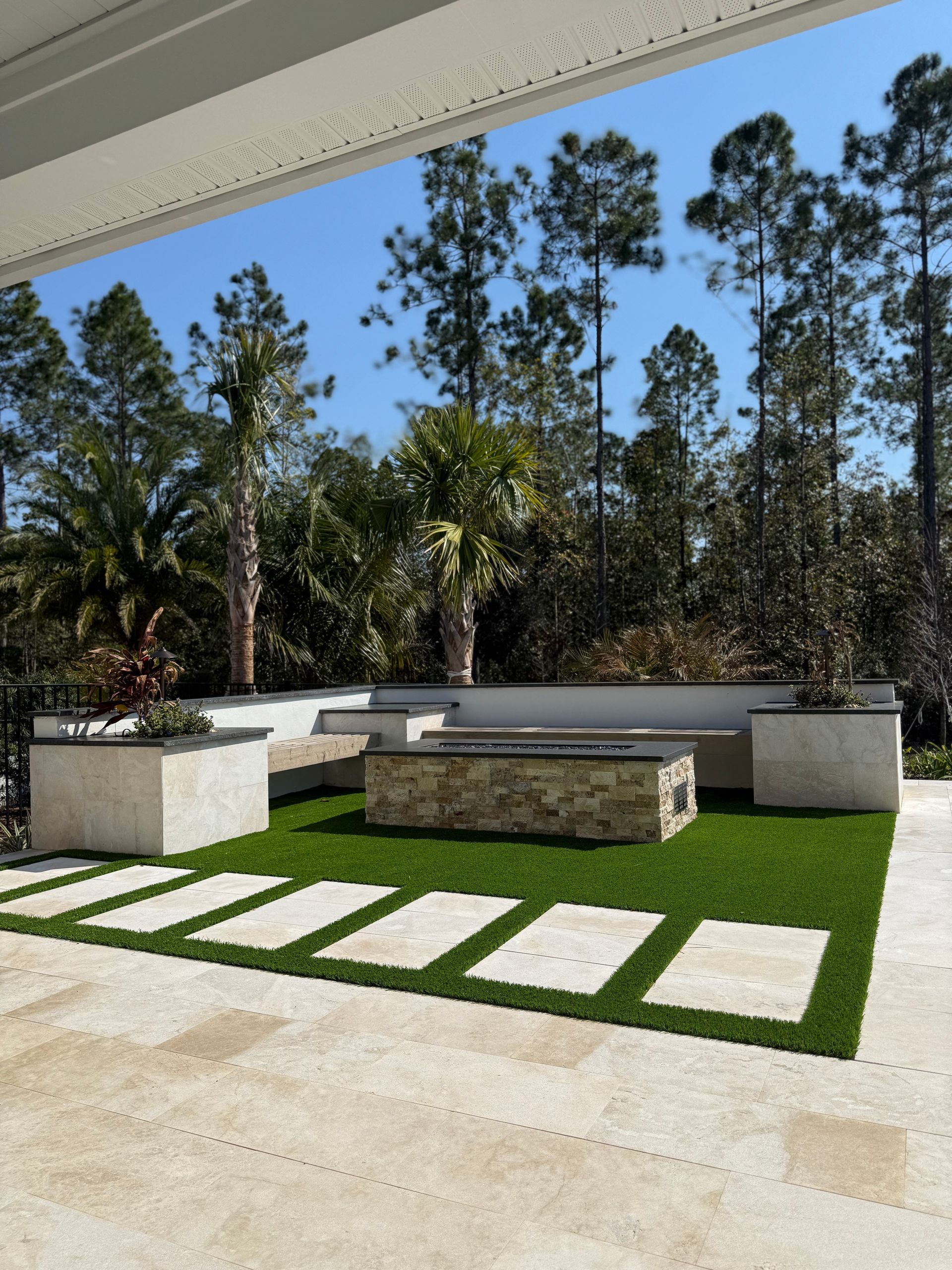 Modern outdoor patio with stone fire pit, green turf, and stepping stone path, trees in background.