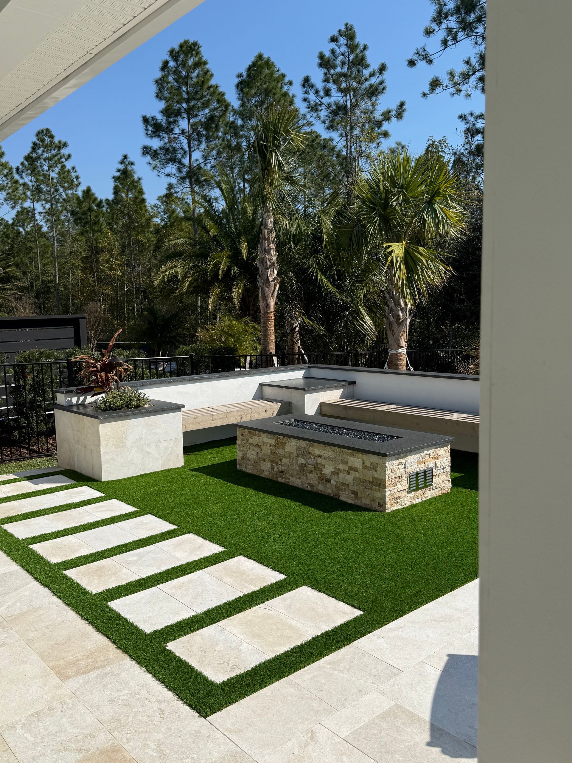 Backyard patio with stone pavers, artificial turf, built-in seating, fire pit, and trees.