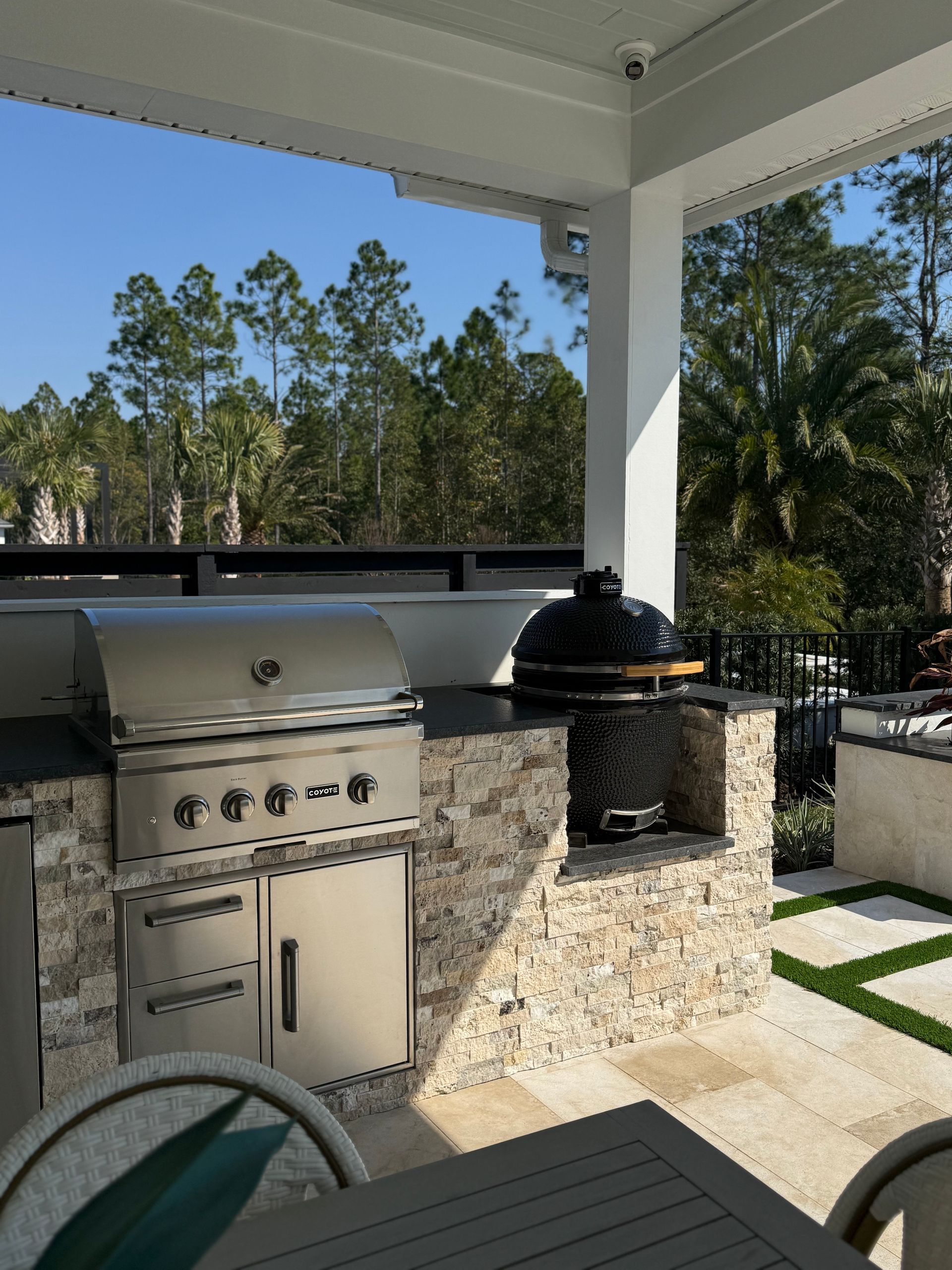Outdoor kitchen with stainless steel grill and kamado-style smoker, stone accents, patio setting.