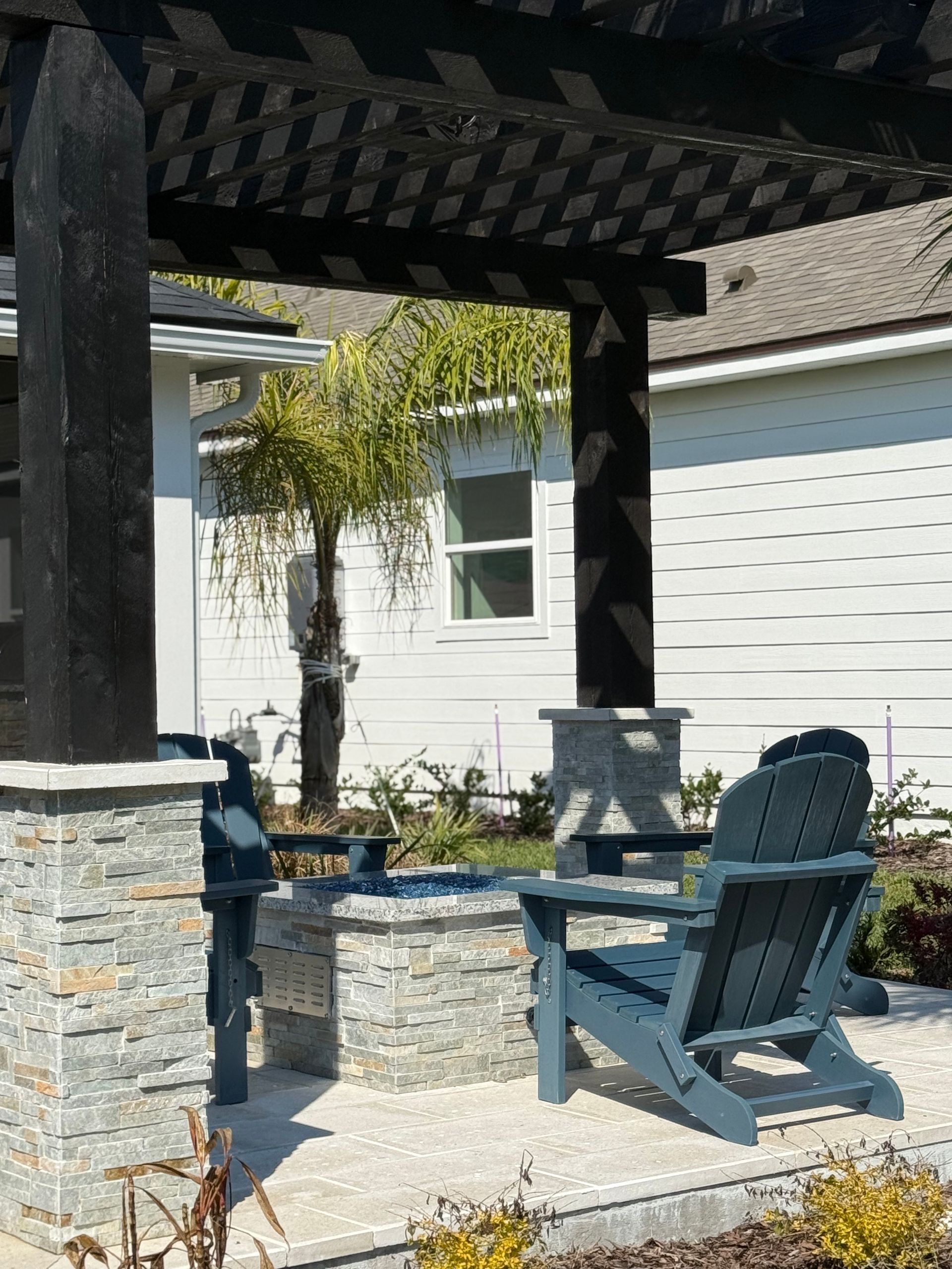A dark blue Adirondack chair sits under a pergola. The structure has dark columns with a stone facade.